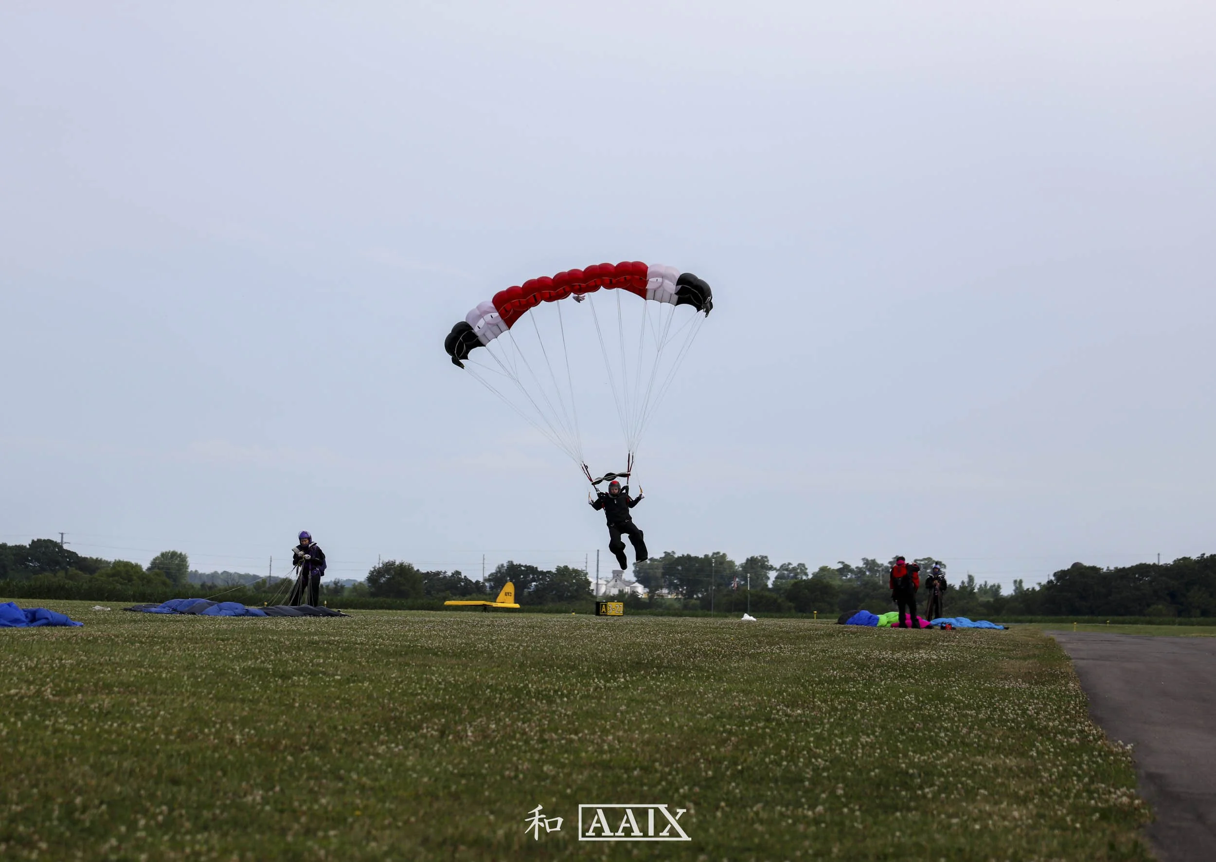 A person parachuting down onto a grassy airstrip with several other people and parachutes on the ground, cloudy sky in the background.