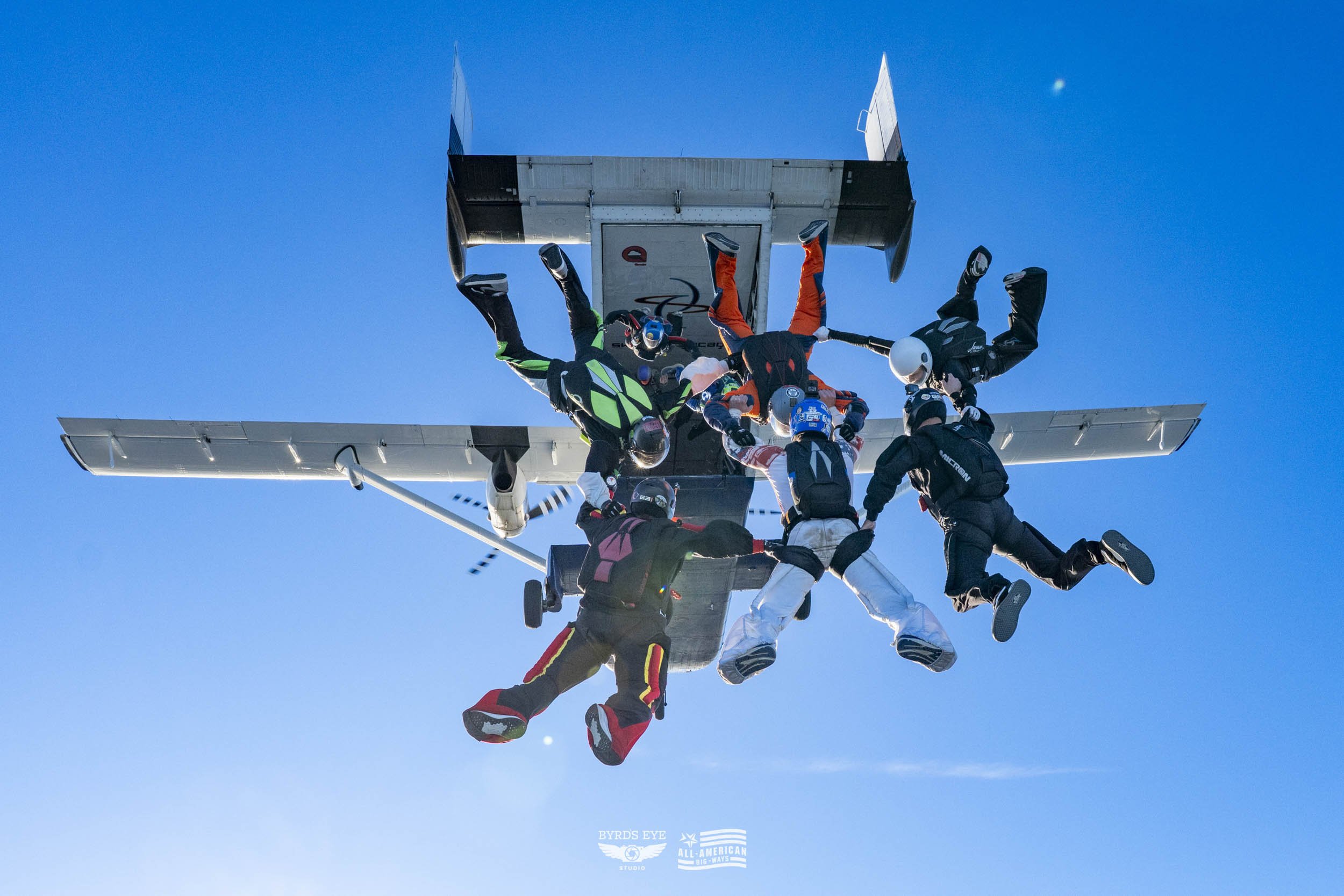 Skydivers mid-air formation around a small aircraft against a blue sky.