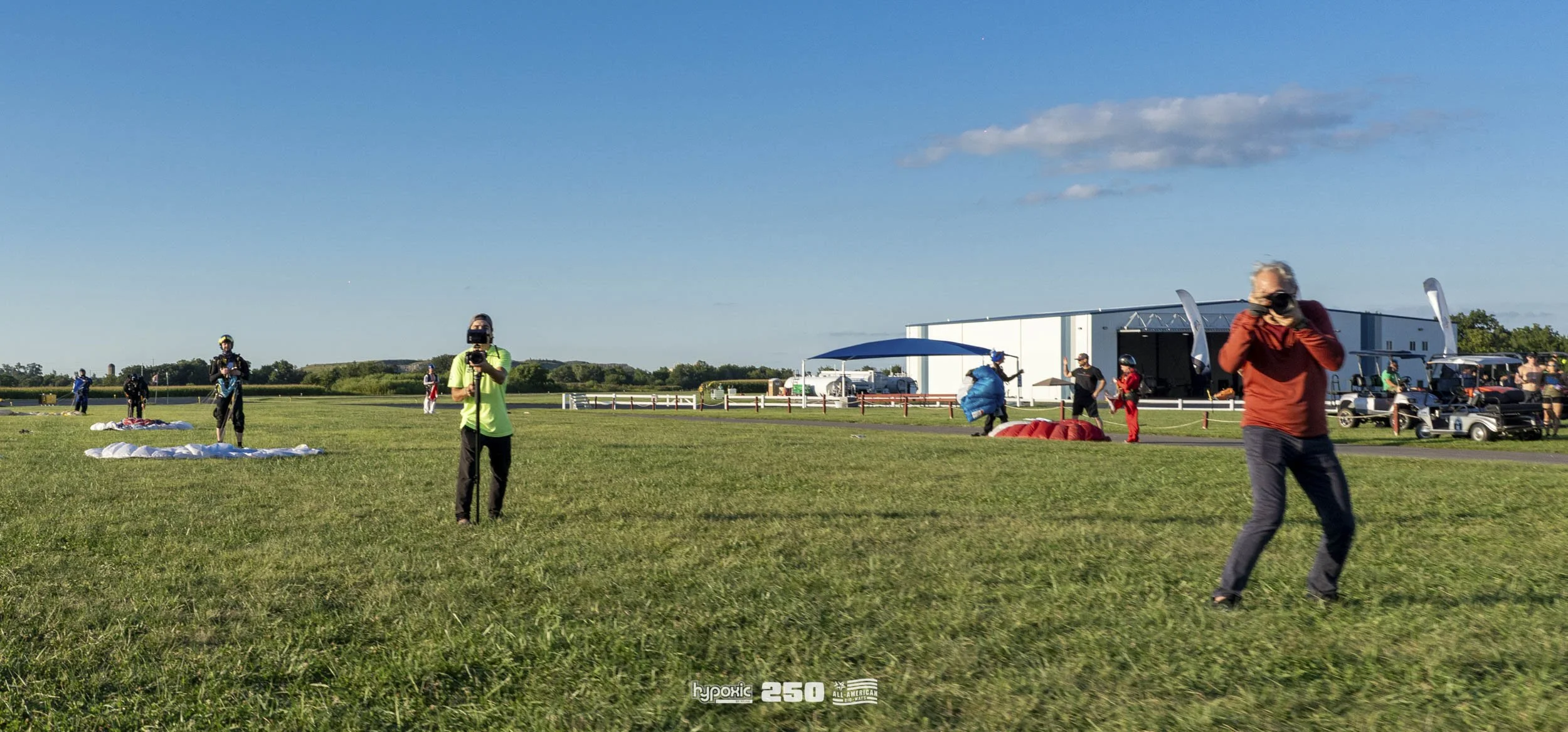 People preparing for a skydiving jump on a grassy field, with parachutes laid out on the ground, and some individuals wearing helmets and gear, during daylight with a clear blue sky.