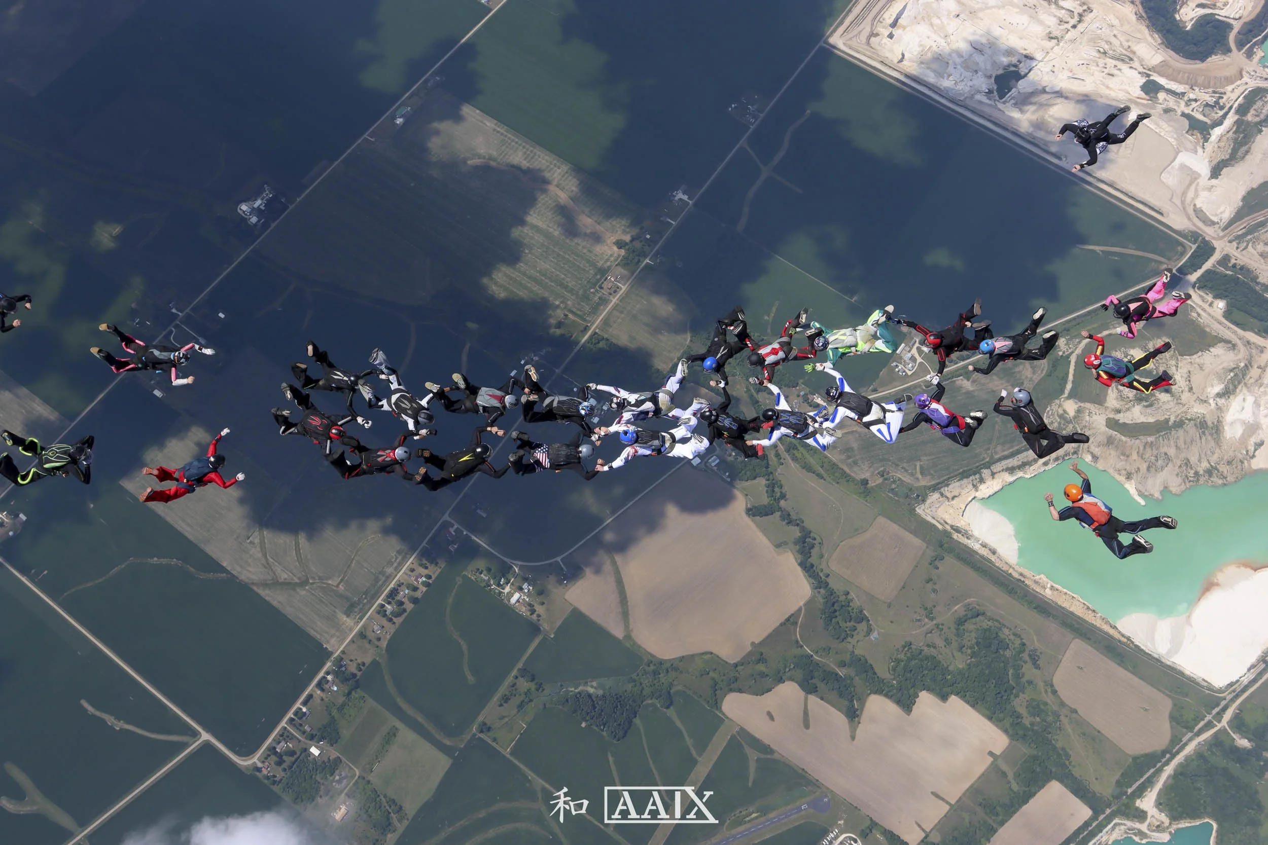 Group of skydivers in freefall, holding hands and forming a line, over farmland and a turquoise lake, with a clear sky above.