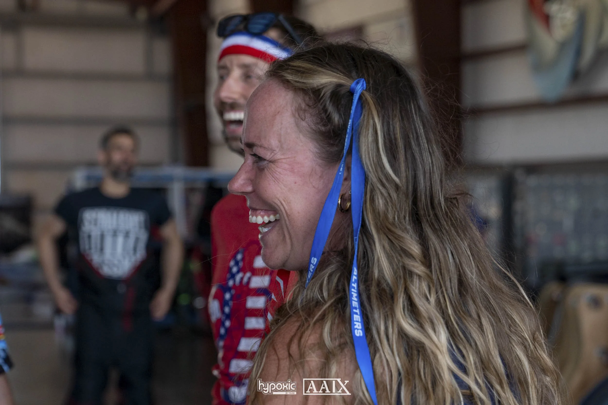 A woman with long, curly blonde hair smiling, wearing a blue ribbon in her hair. Behind her, a man in a red, white, and blue headband and another person in a black shirt with white text, in a room with wooden walls.