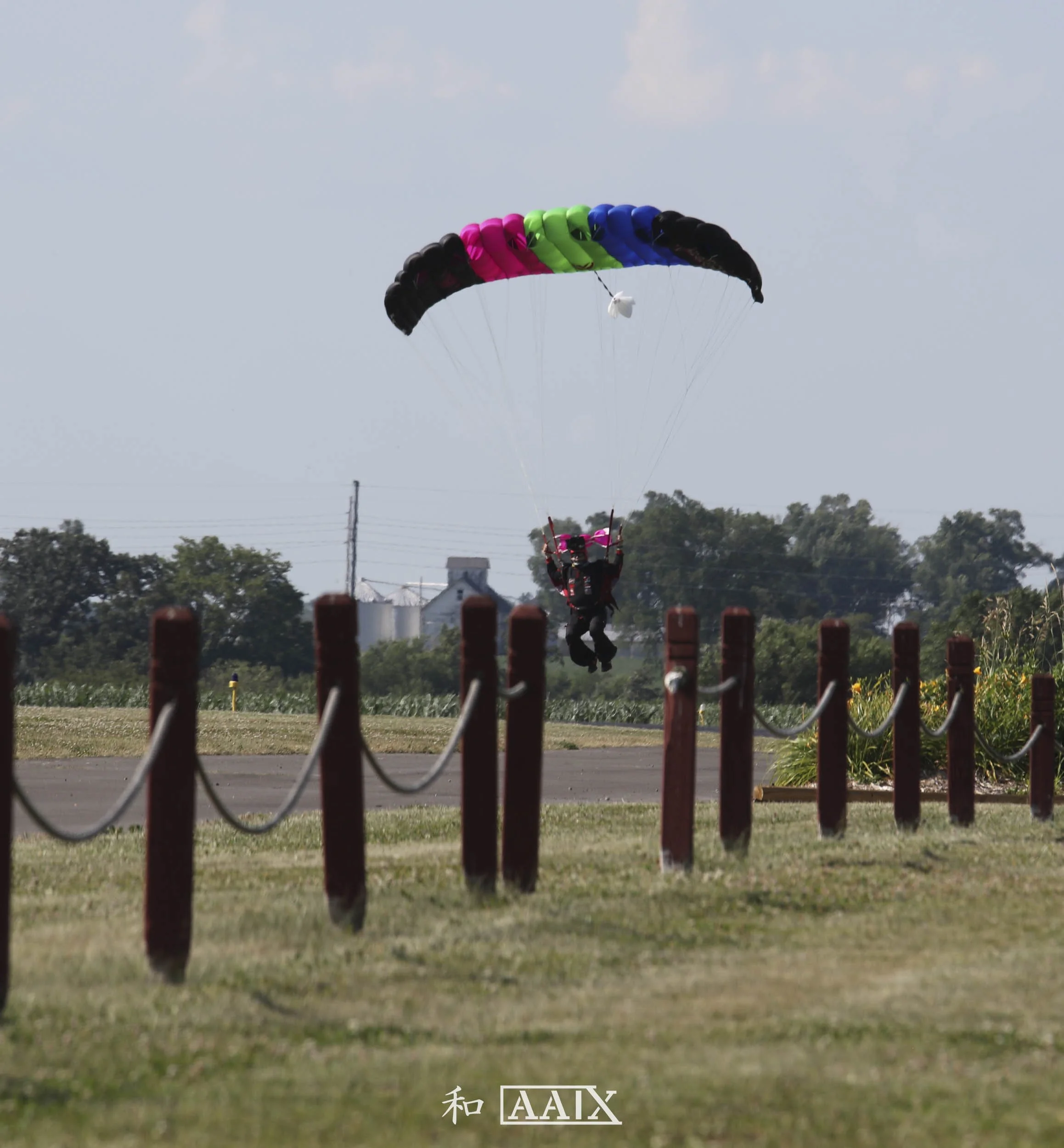 A person parachuting with a black, pink, green, and blue parachute over a grassy field, with a clear sky and trees in the background.