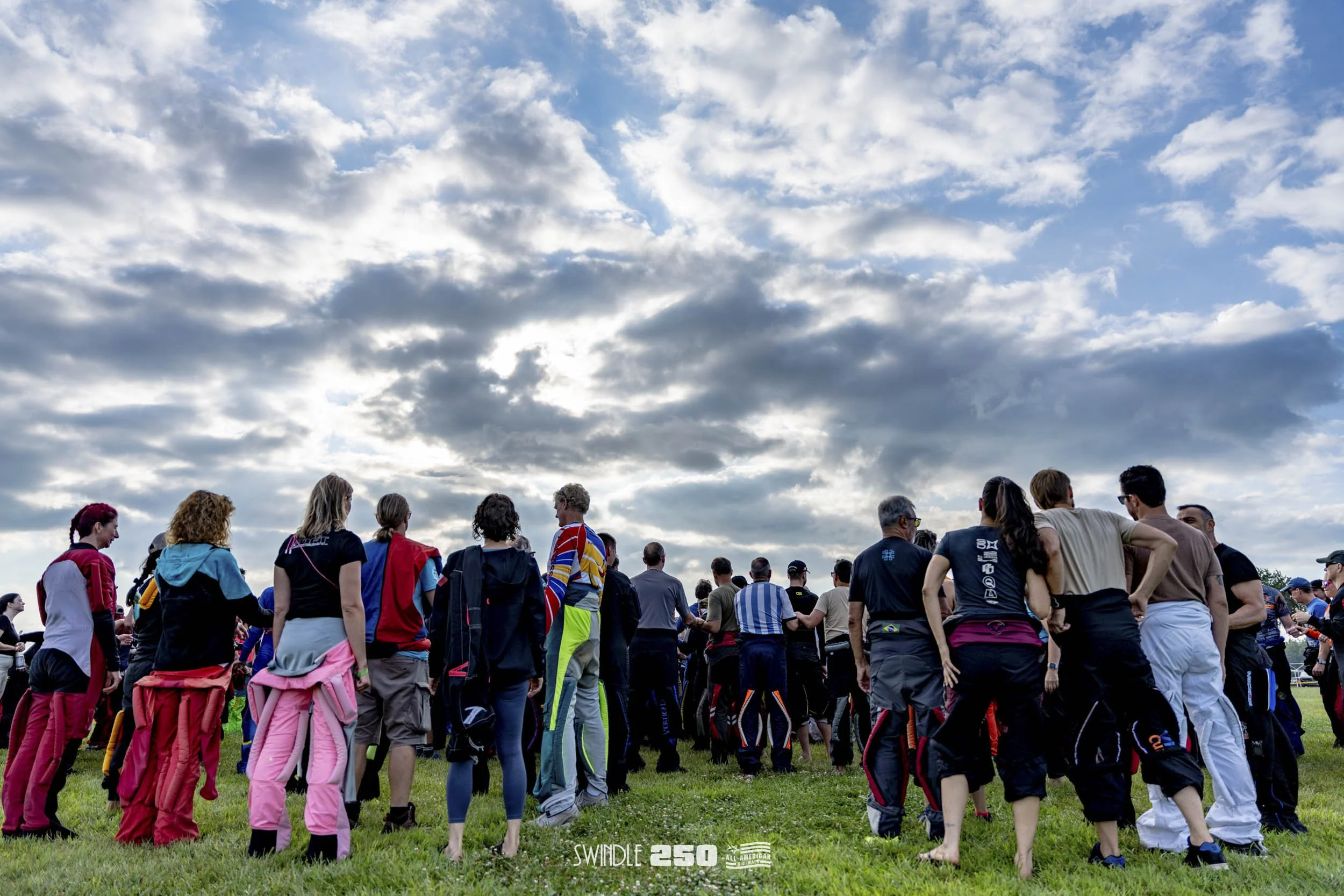 A group of people gathered outdoors on a grassy field under a blue sky with clouds, some wearing racing suits and gear, possibly for an event or race.
