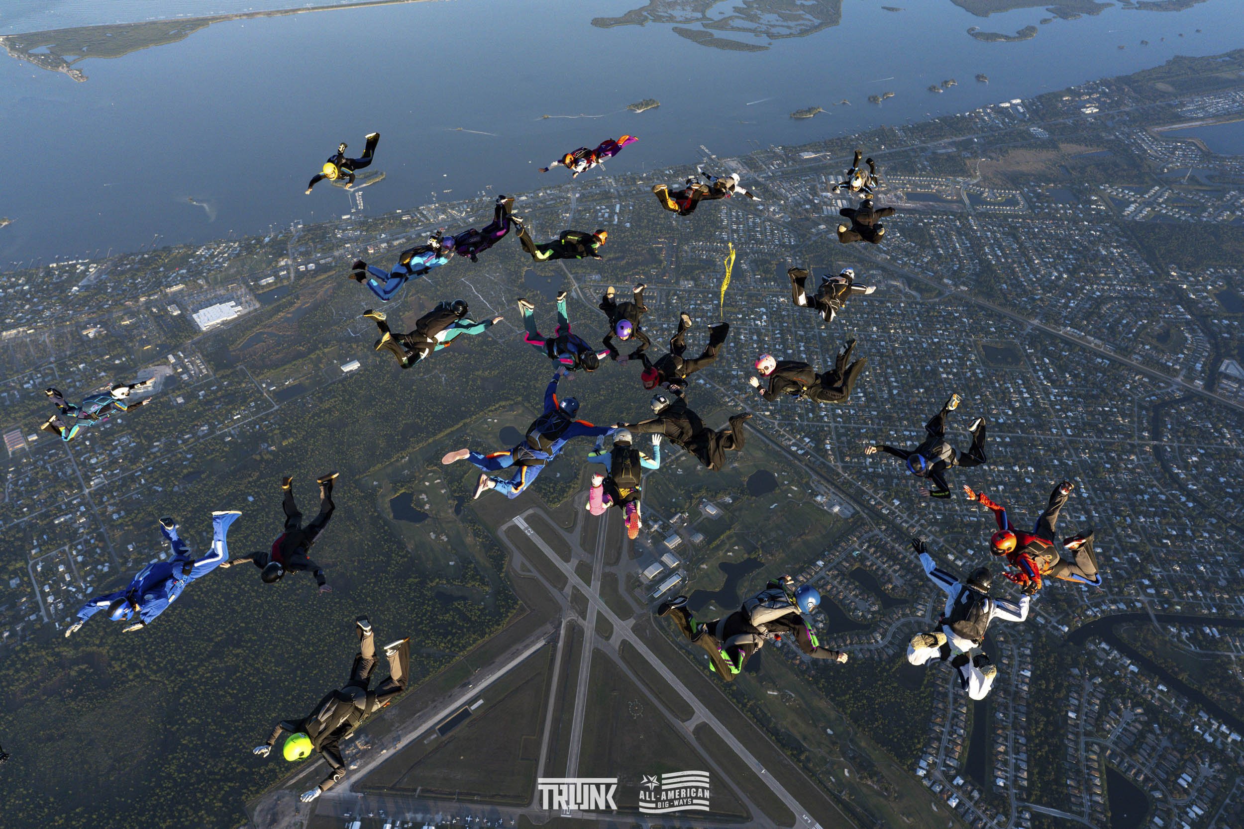 Group of skydivers in free fall above a landscape with a river, city, and wooded areas.