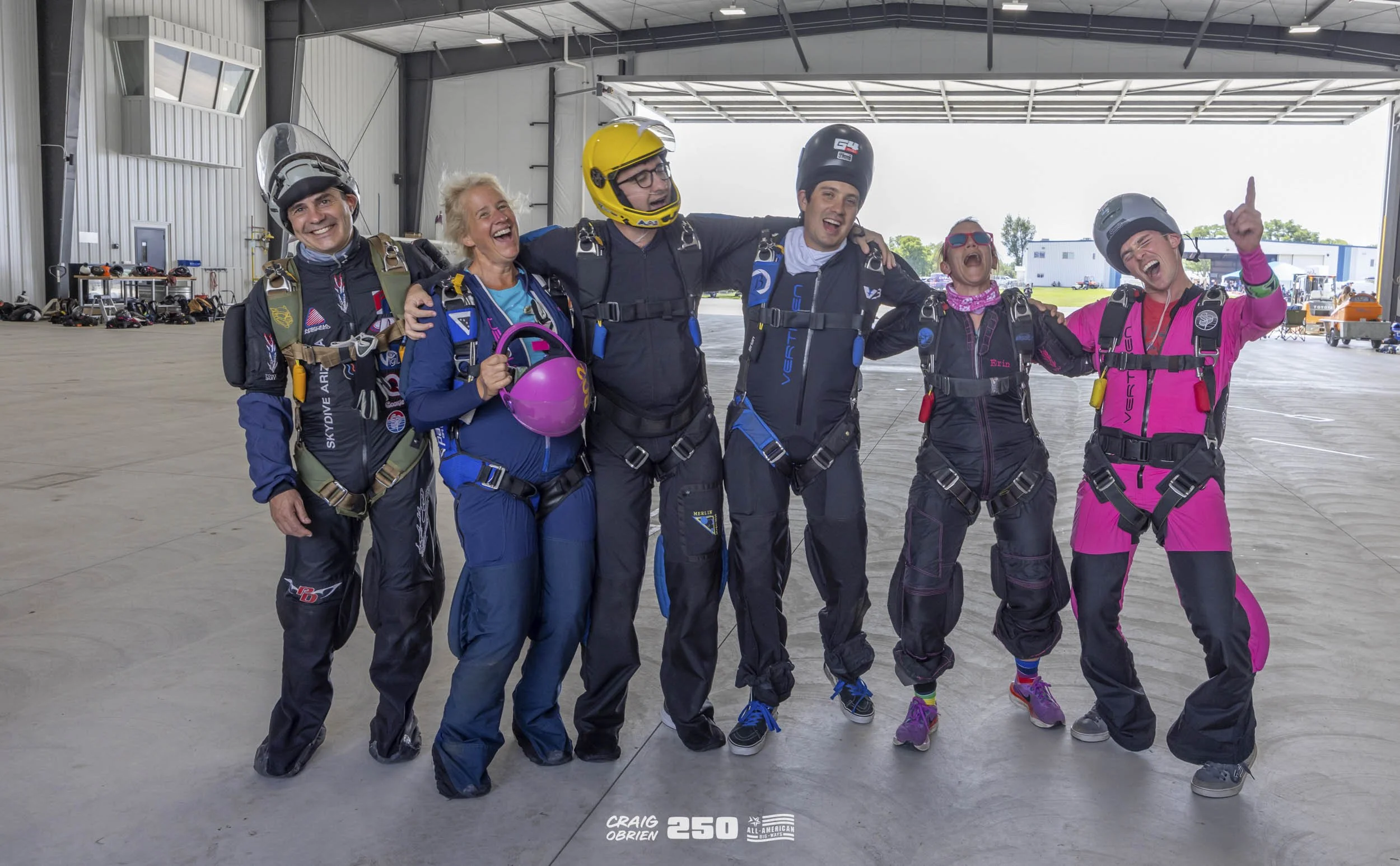 Group of skydivers in jumpsuits and helmets celebrating inside an aircraft hangar.
