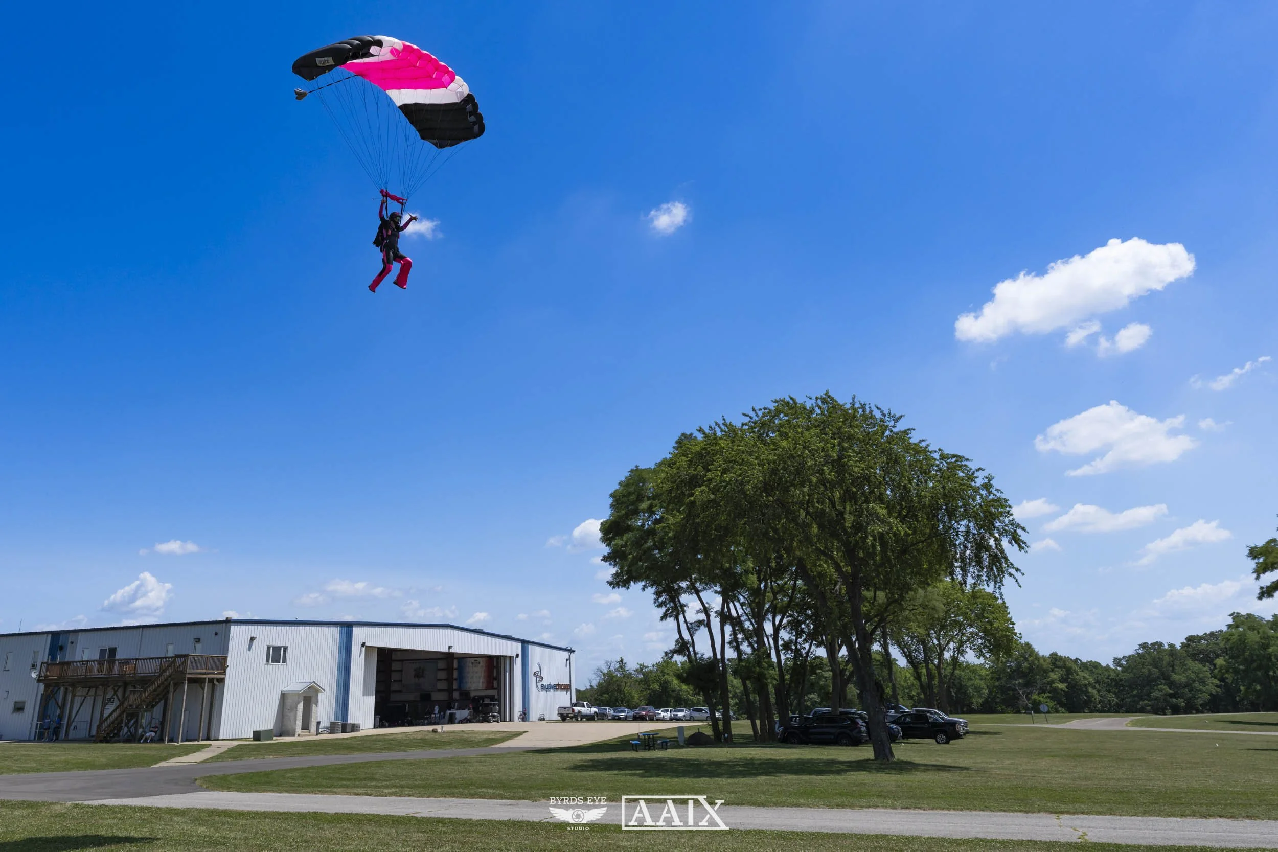 Person skydiving with a pink, white, and black parachute above a grassy field and a large white hangar building with parked cars, trees, and a blue sky with scattered clouds.