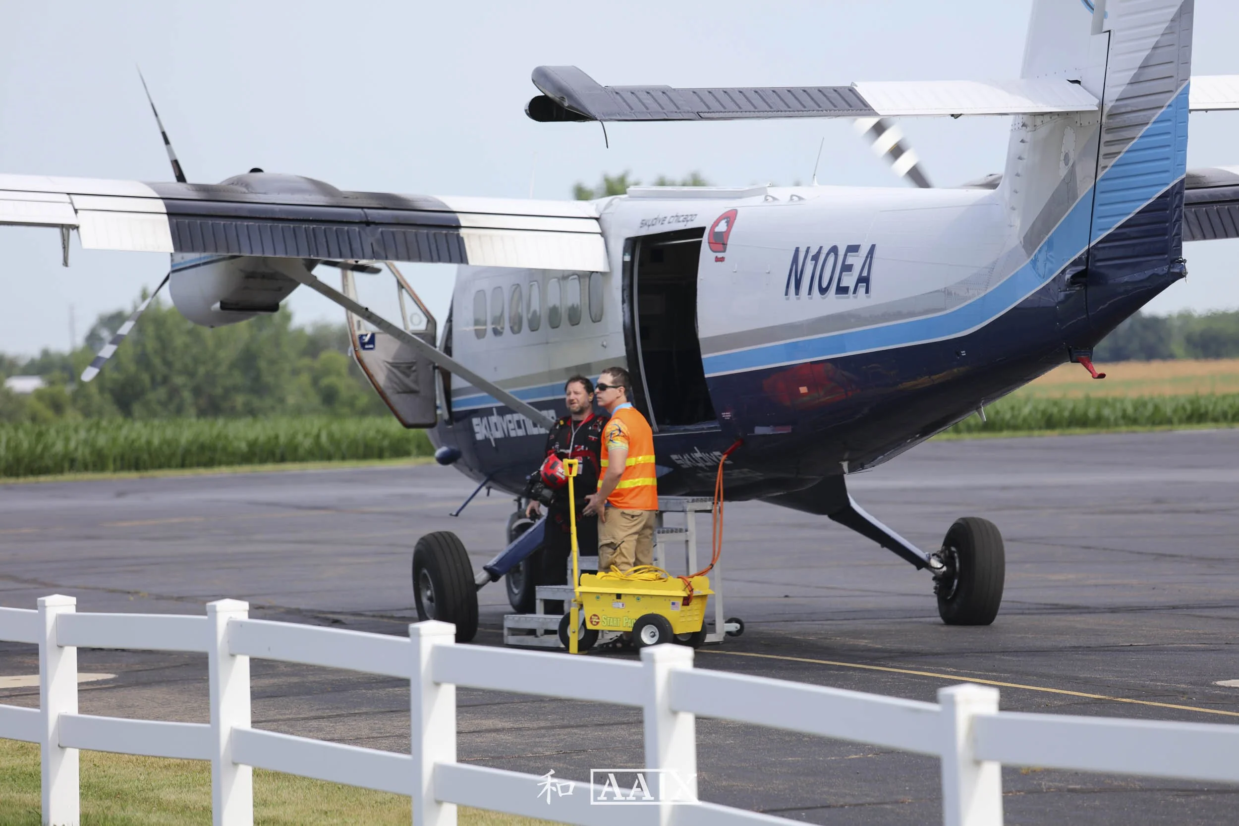 Two men standing outside a small aircraft on the tarmac, one in a black flight suit and the other in an orange safety vest, with a yellow emergency response cart nearby.