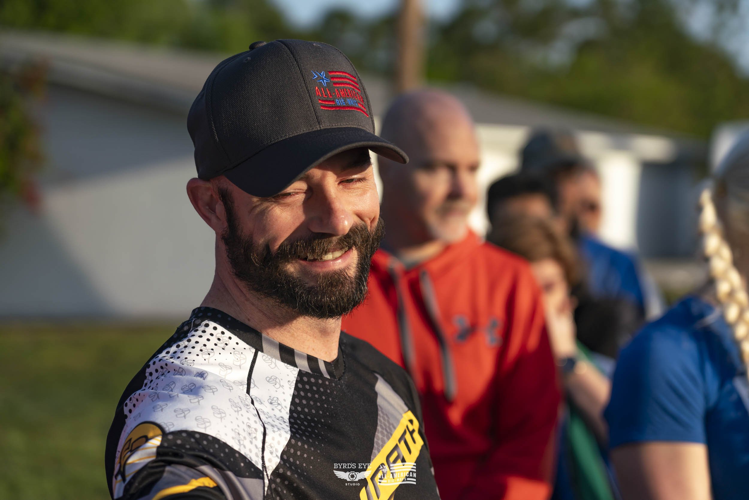 A smiling man wearing a black cap and cycling jersey standing outdoors among a group of people backlit by sunlight.