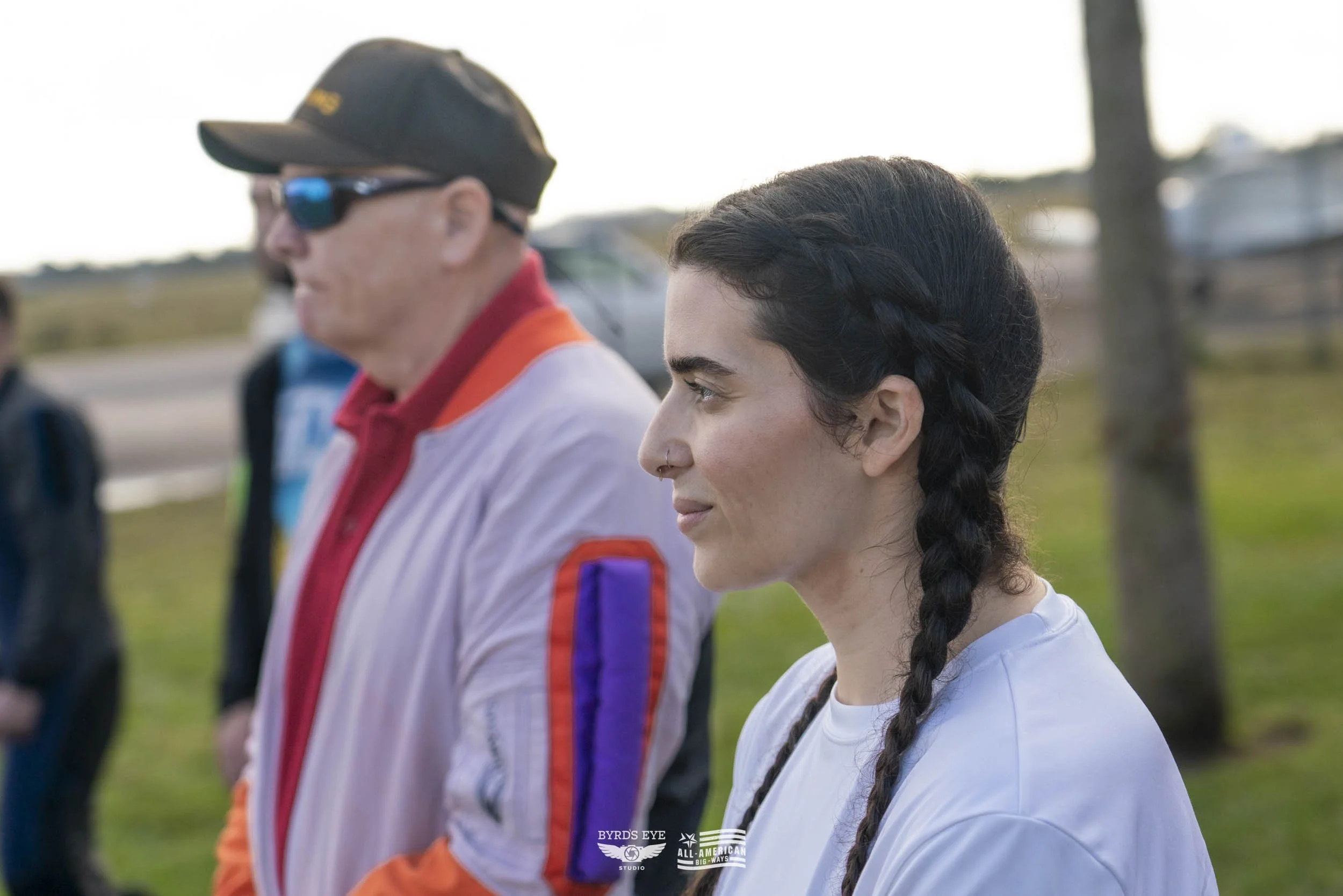 A young woman with dark braided hair and a nose ring stands outdoors in profile, wearing a white shirt, with a man in sunglasses and racing jacket beside her, both facing right, in a grassy area with trees.