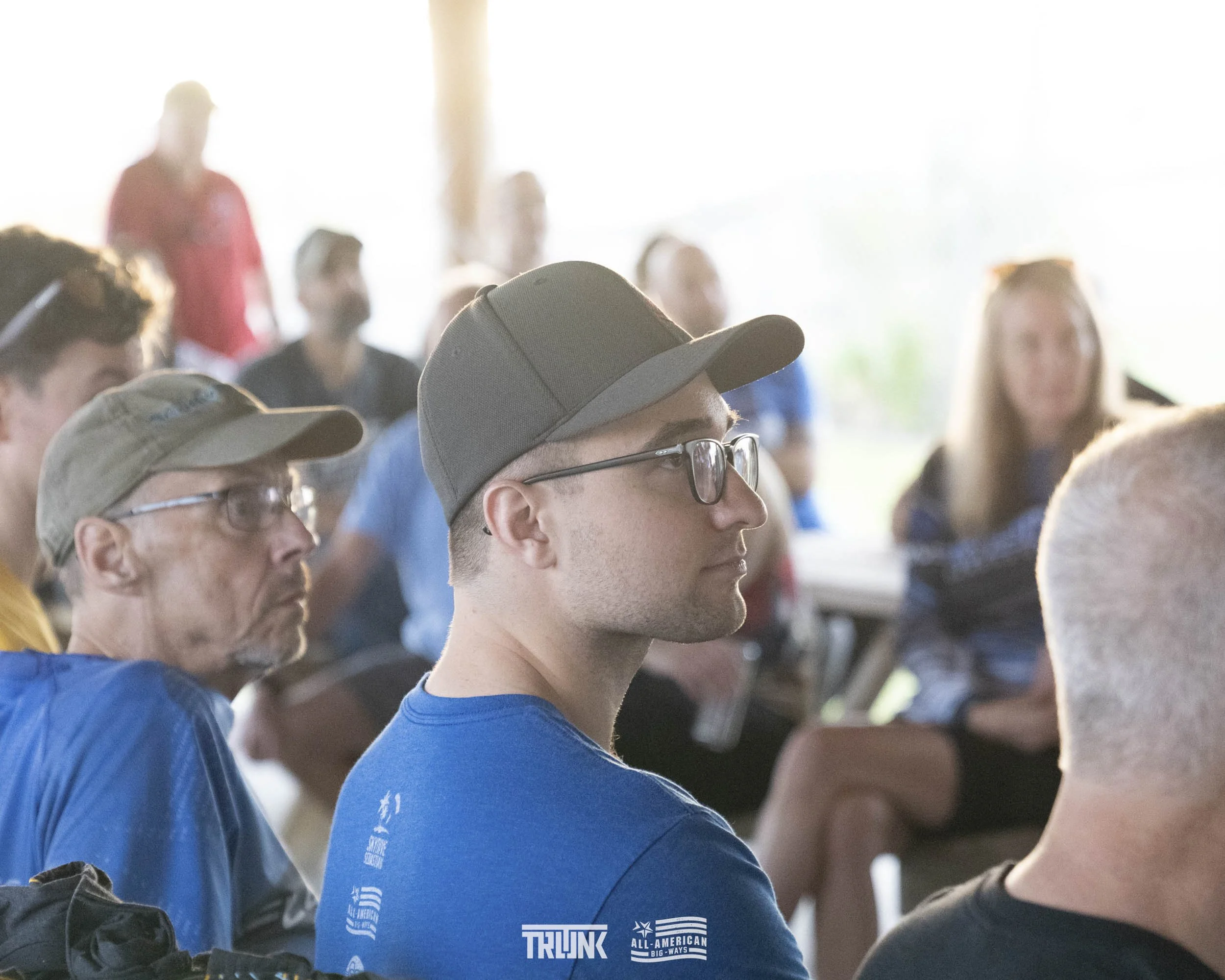 A group of people sitting indoors, attentively listening, with some wearing glasses and caps, and a bright background with natural light.