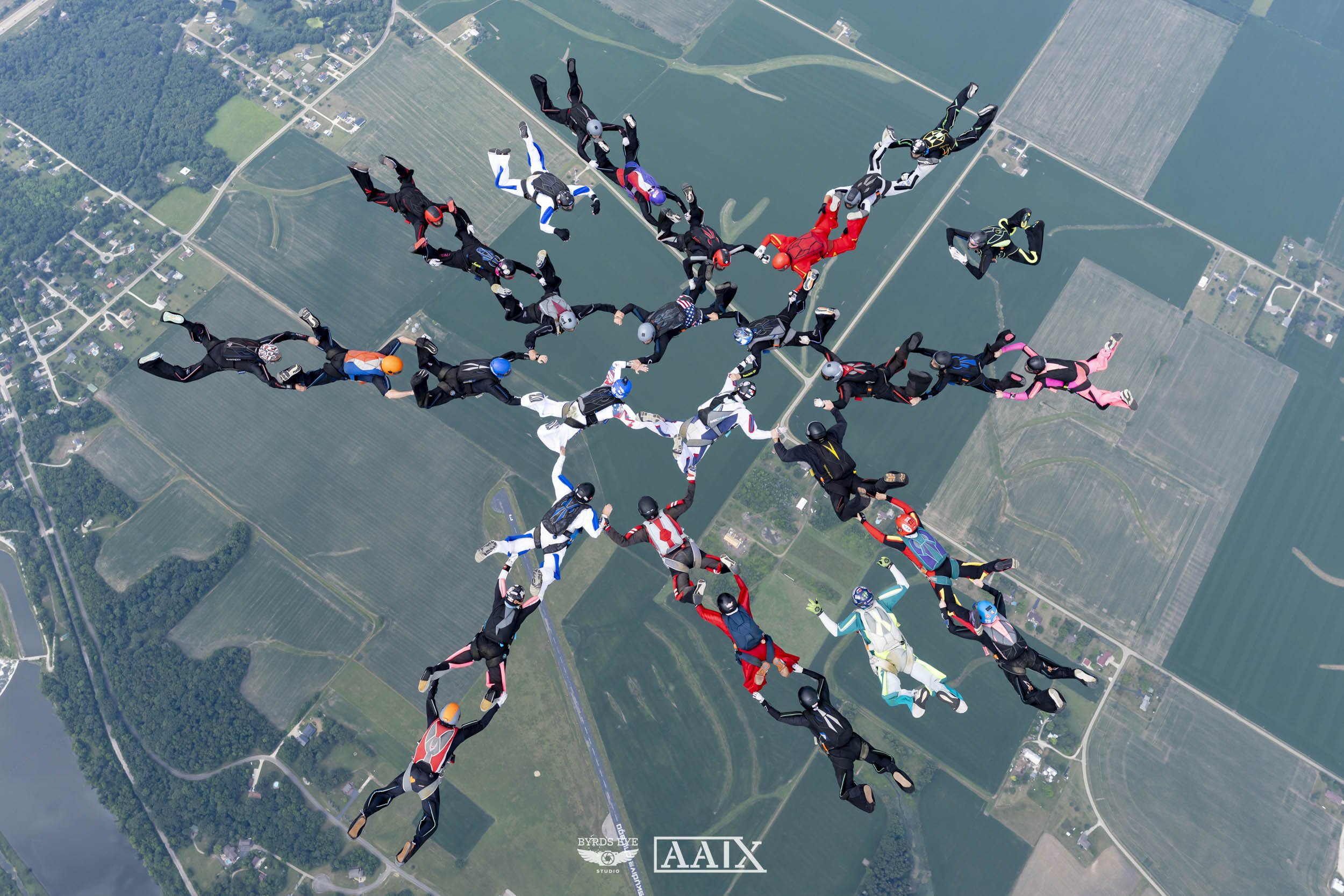 A group of skydivers in jumpsuits forming a star shape while free-falling over farmland and bodies of water.