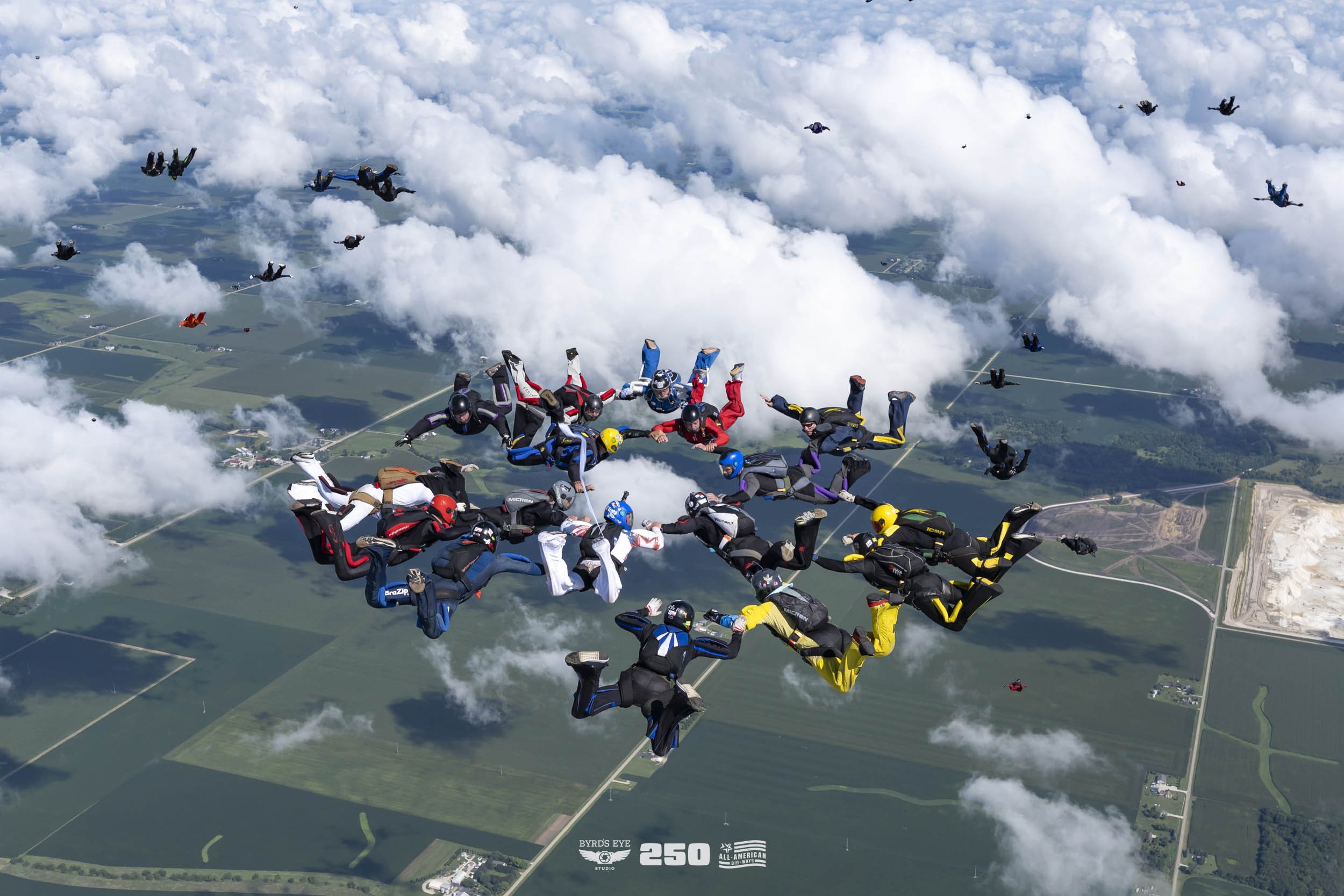 Group of skydivers forming a heart shape in the sky above farmland, with additional skydivers free-falling in the background.