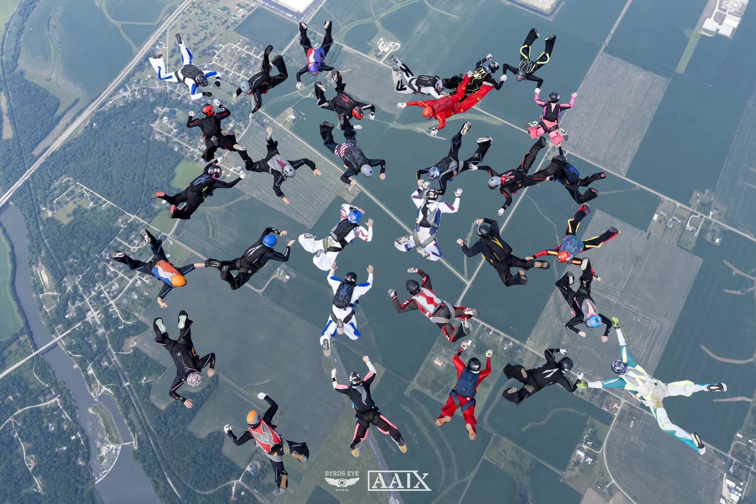 Group of 29 skydivers in colorful jumpsuits and helmets free-falling in a circular formation over farmland and a river, with fields and roads below.