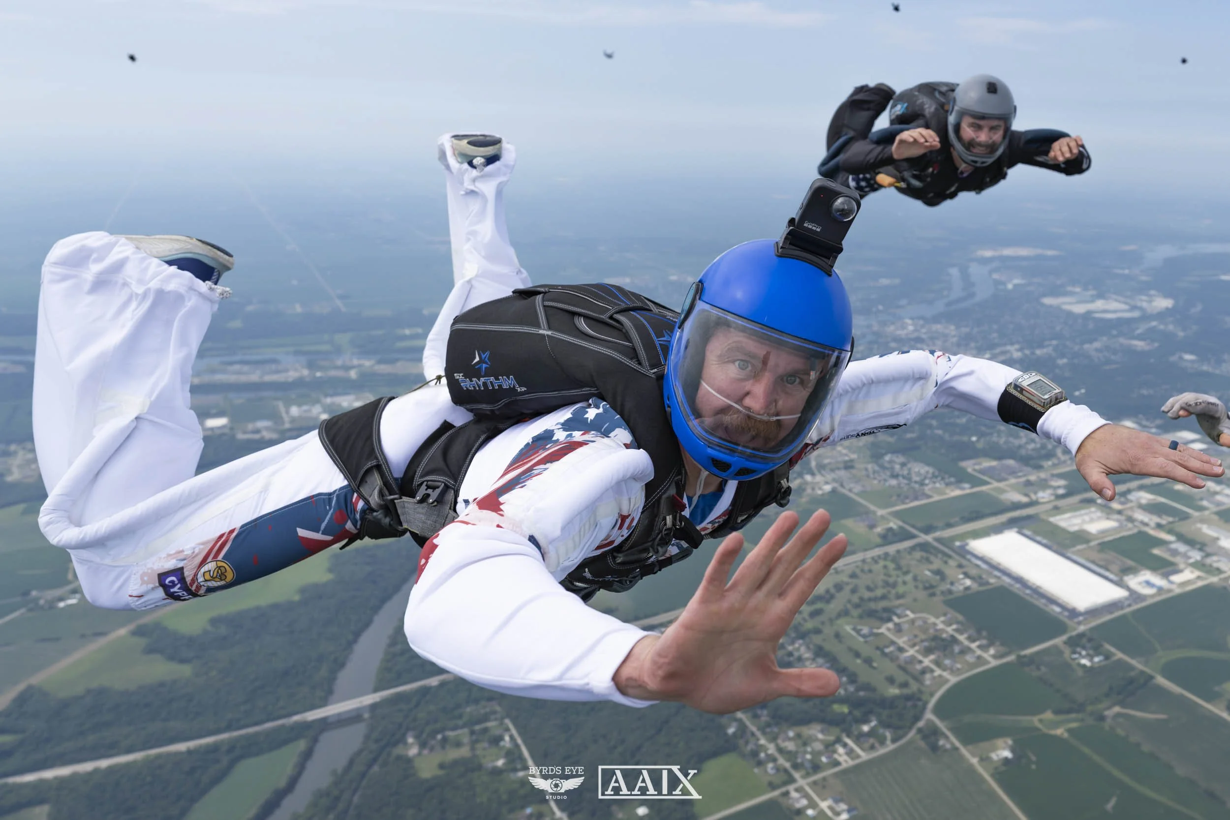 Two skydivers free-falling above a landscape, one in the foreground with a blue helmet and a GoPro, and the other in the background with a gray helmet, both wearing jumpsuits and gear, with fields and a river below.