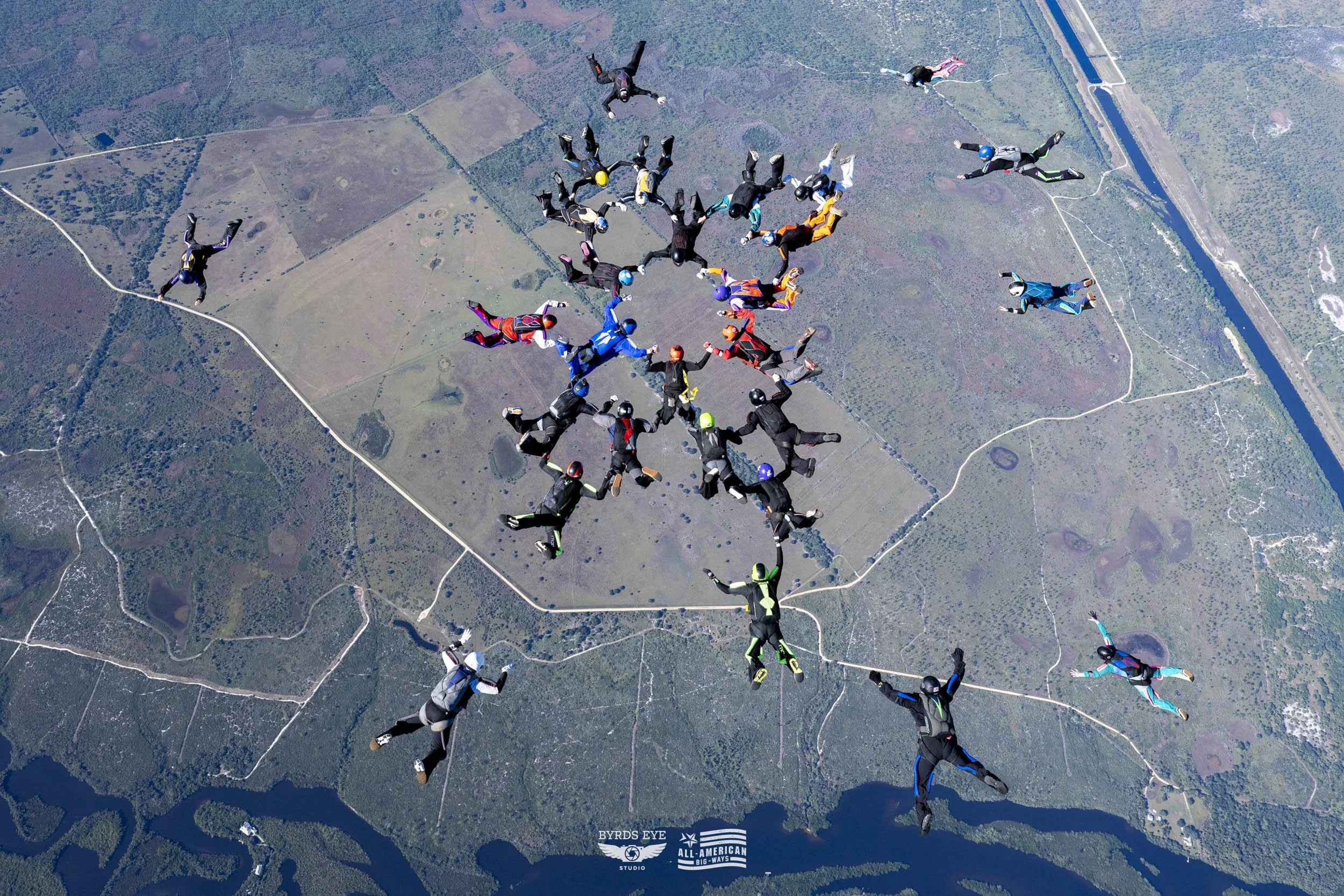 Group of skydivers in free fall formation over a landscape with fields, roads, and water bodies.