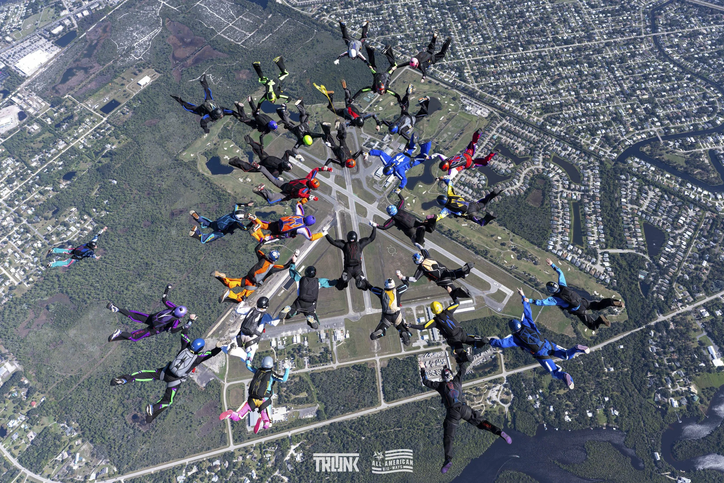 Hundreds of skydivers in colorful jumpsuits and helmets are performing a synchronized group skydiving formation over a cityscape with a river, roads, and buildings below.