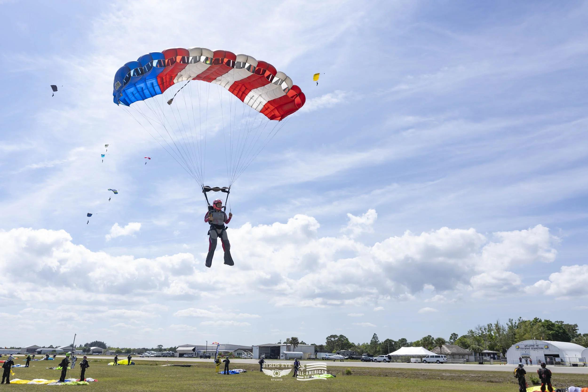 A person parachuting with a red, white, and blue parachute during a skydiving event at an airfield, with other parachutes in the sky and people on the ground.