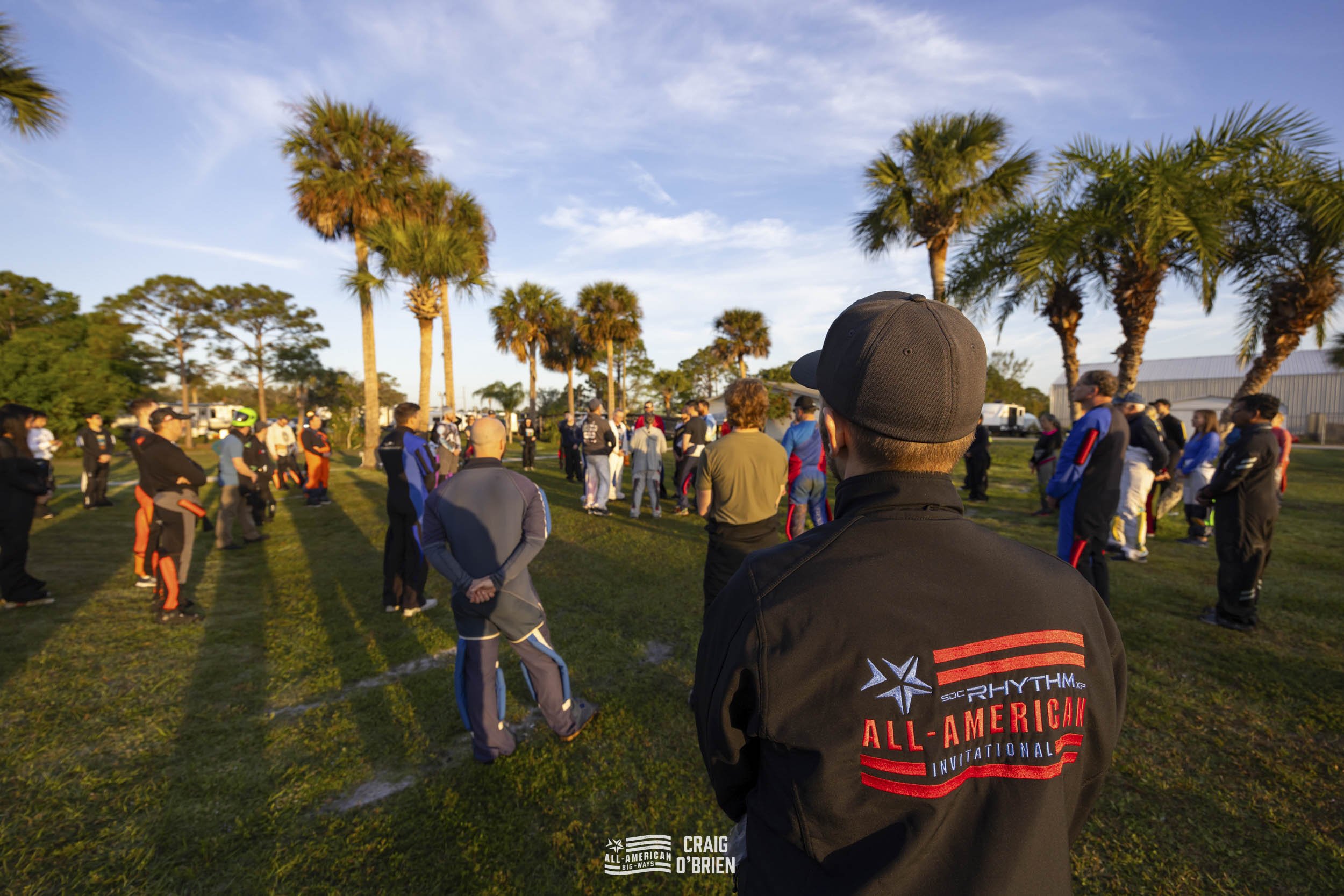 Group of people gathering outdoors on grassy field with palm trees, social distancing, in the late afternoon or early evening sunlight.