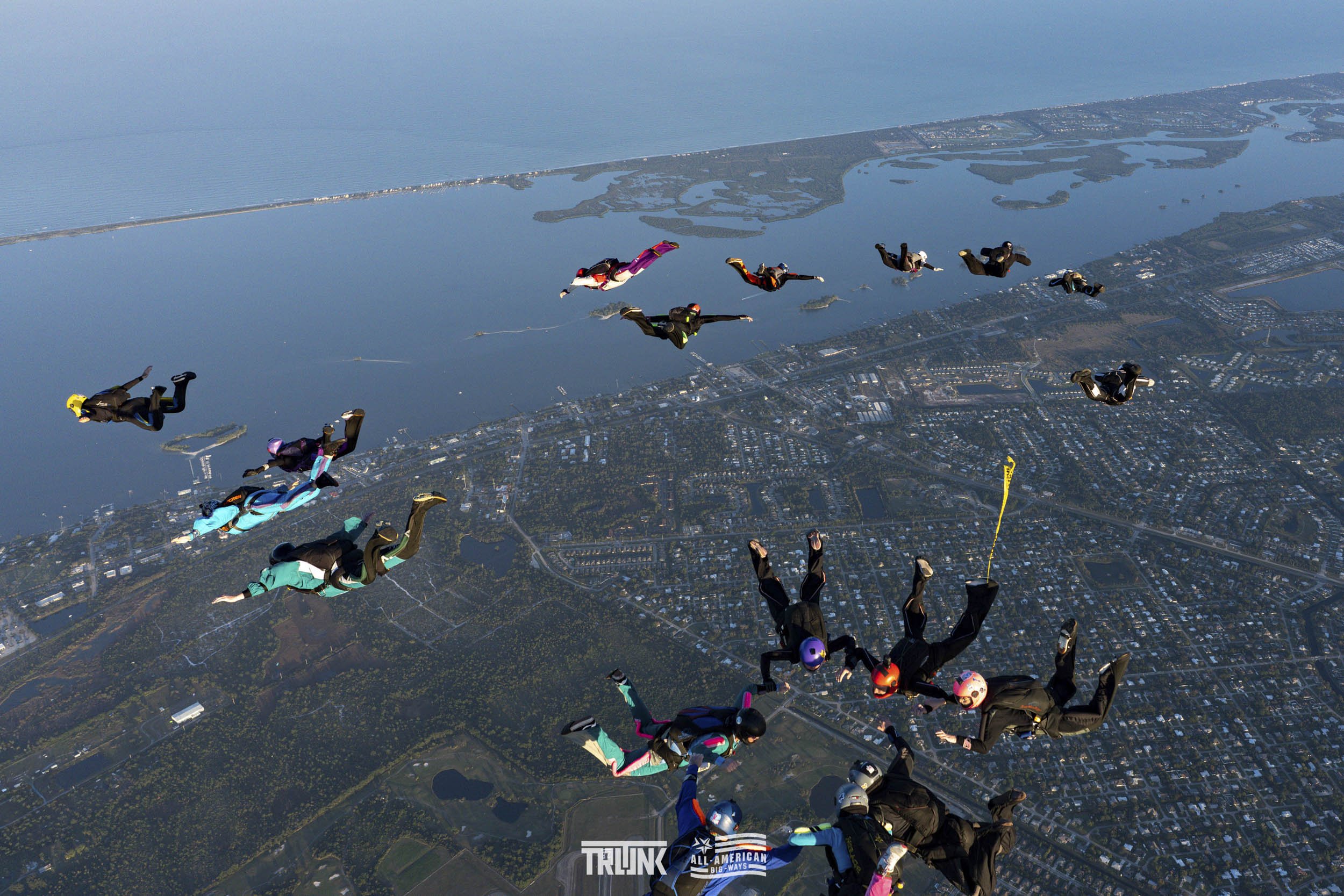 Group of skydivers in free fall over a coastal landscape with water, islands, and a city underneath on a clear day.