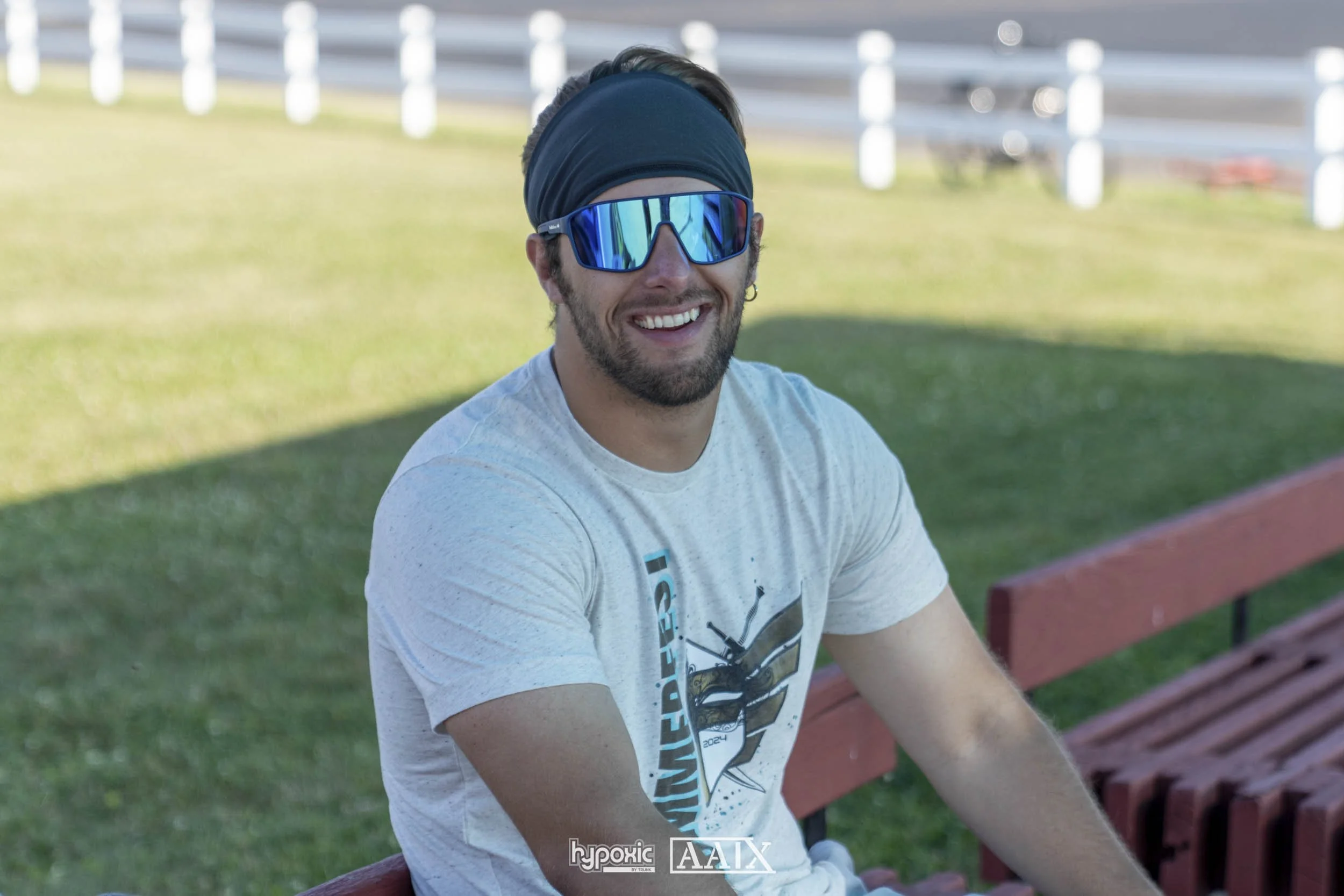 A man sitting on a park bench outdoors, wearing sunglasses, a headband, and a light-colored t-shirt, smiling at the camera.