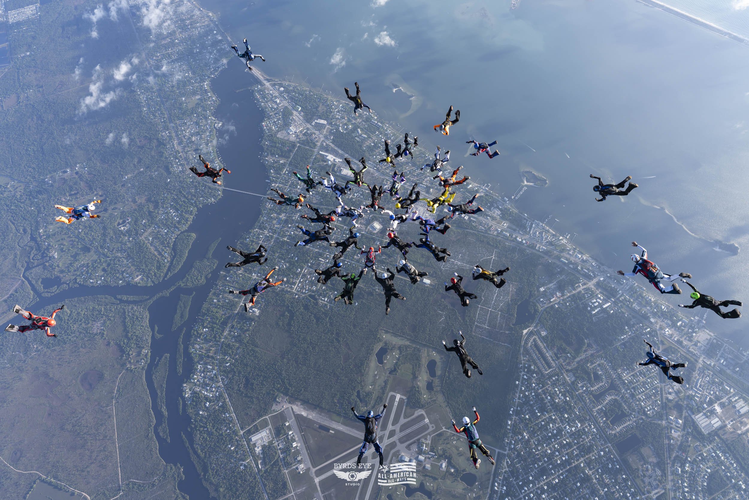 Skydivers in free fall over a city and water body, with a visible airport runway below.