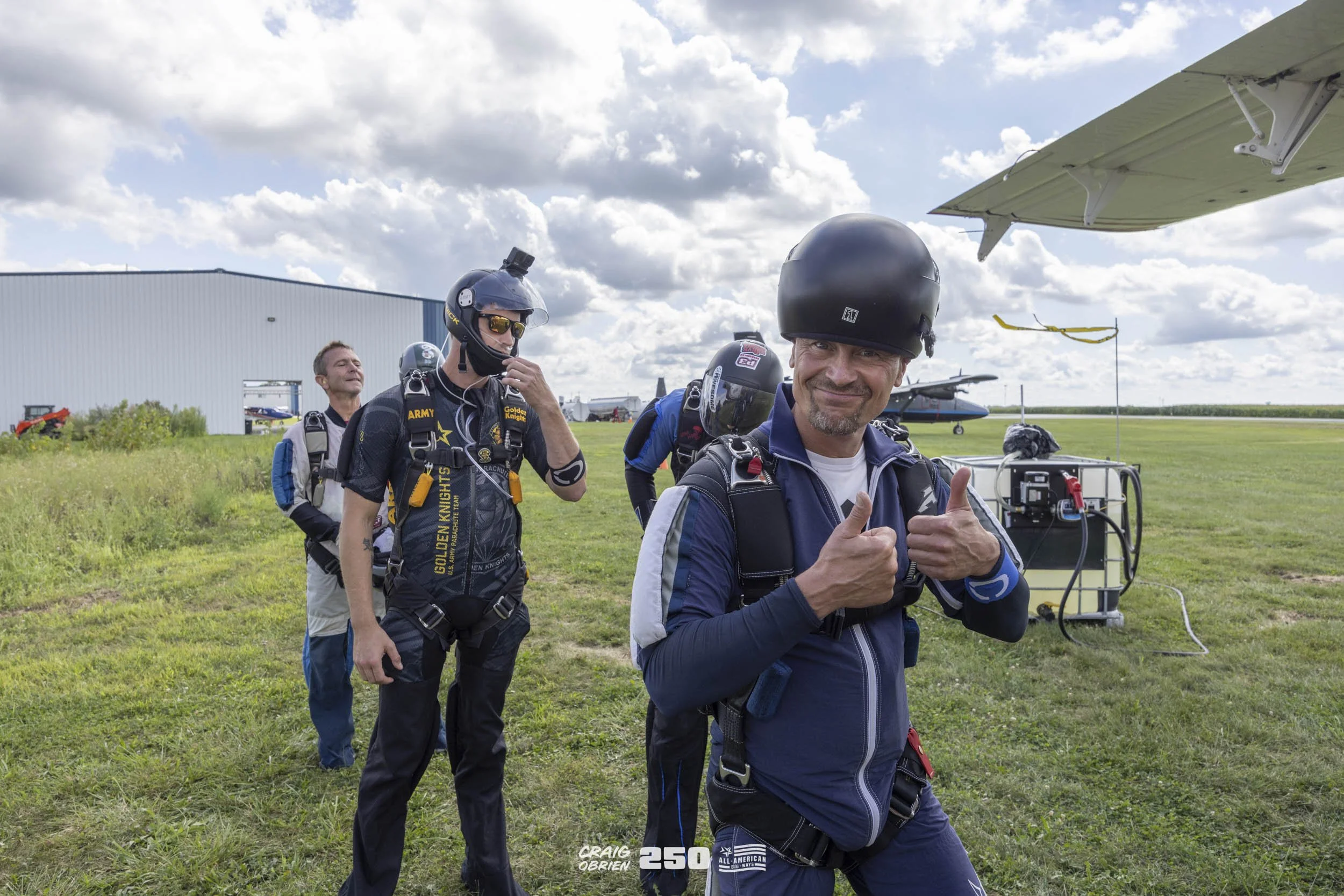 A group of skydivers preparing for a jump on a grassy airfield, with airplanes in the background, one man smiling and giving a thumbs-up while wearing a helmet and skydiving gear.