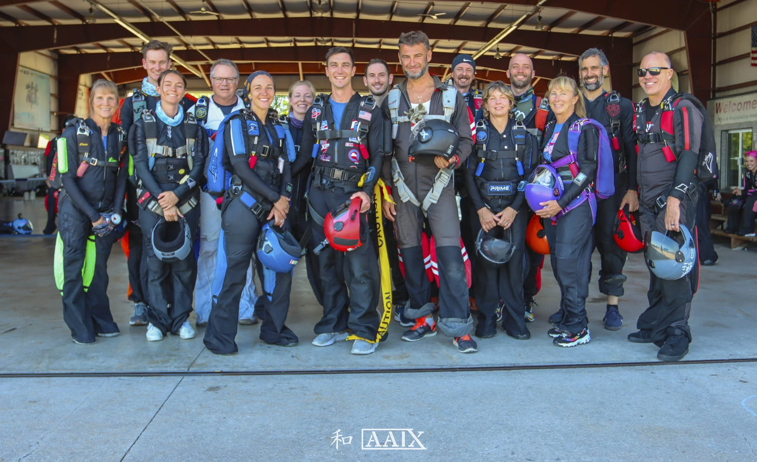 Group of skydivers in jumpsuits and helmets, standing together with harnesses, inside a hangar.
