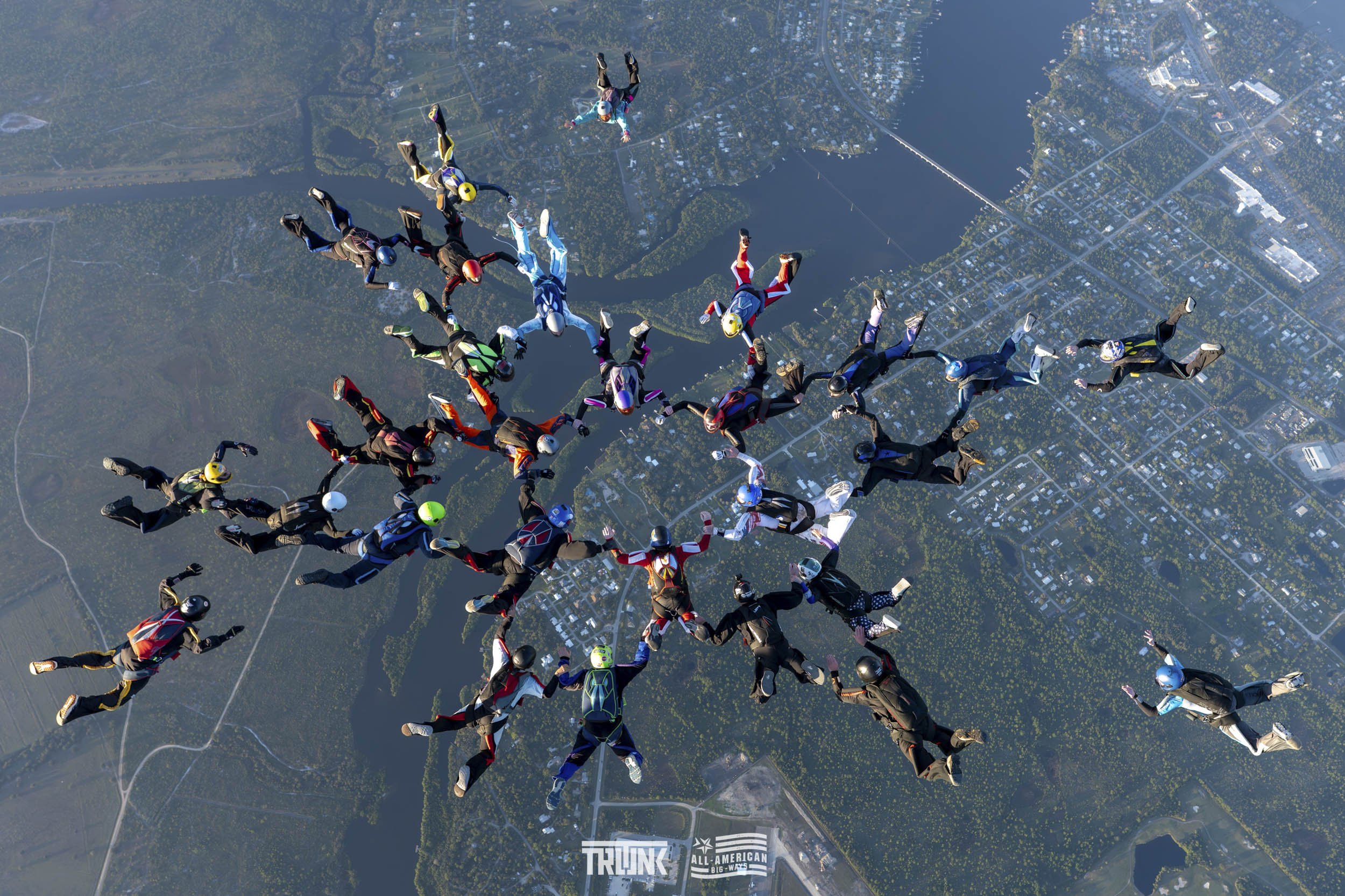 Group of skydivers in colorful jumpsuits and helmets free-falling together over a landscape with water and roads.