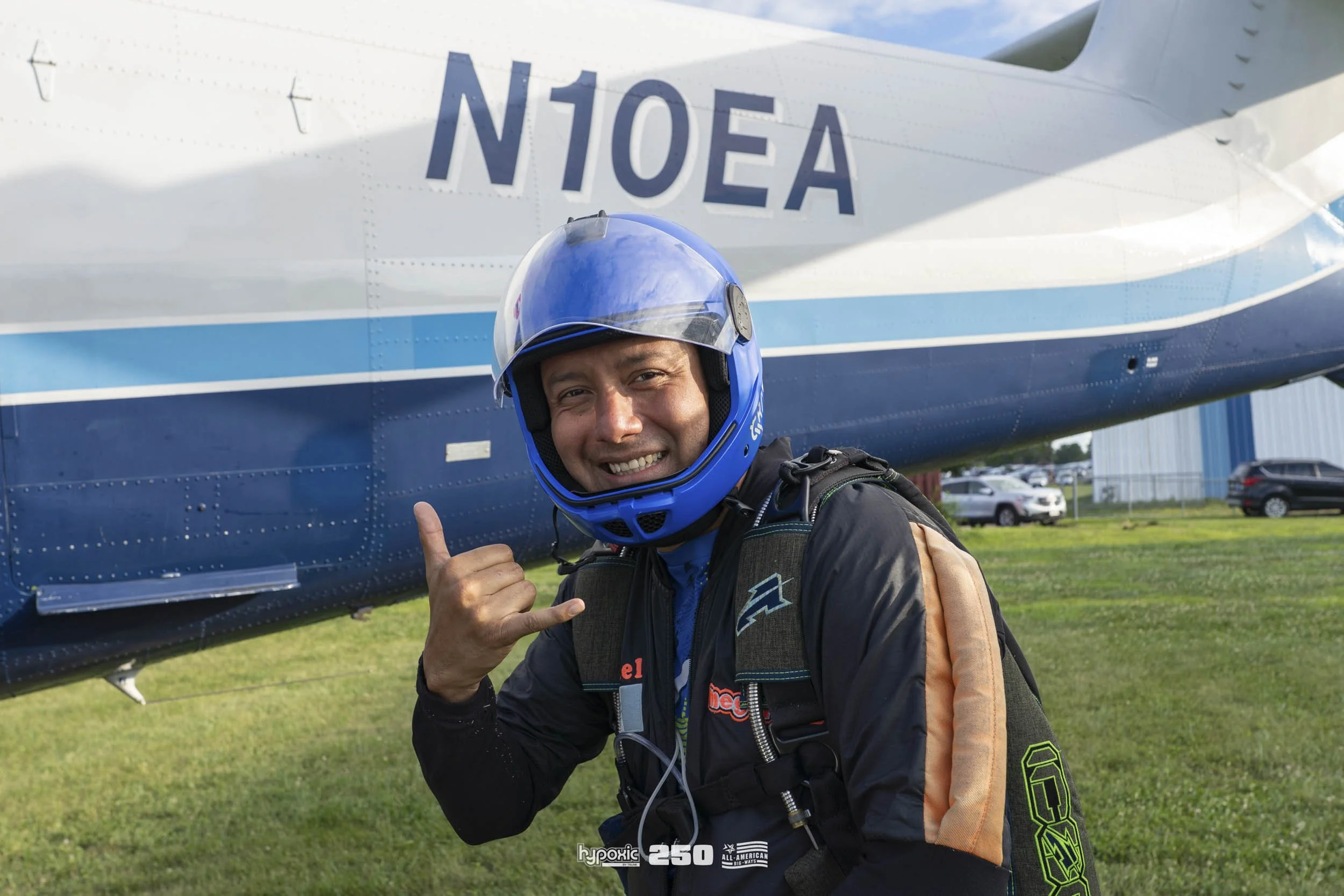 A smiling man in a blue helmet and black jumpsuit making the shaka sign in front of a helicopter with the registration N10EA on it.