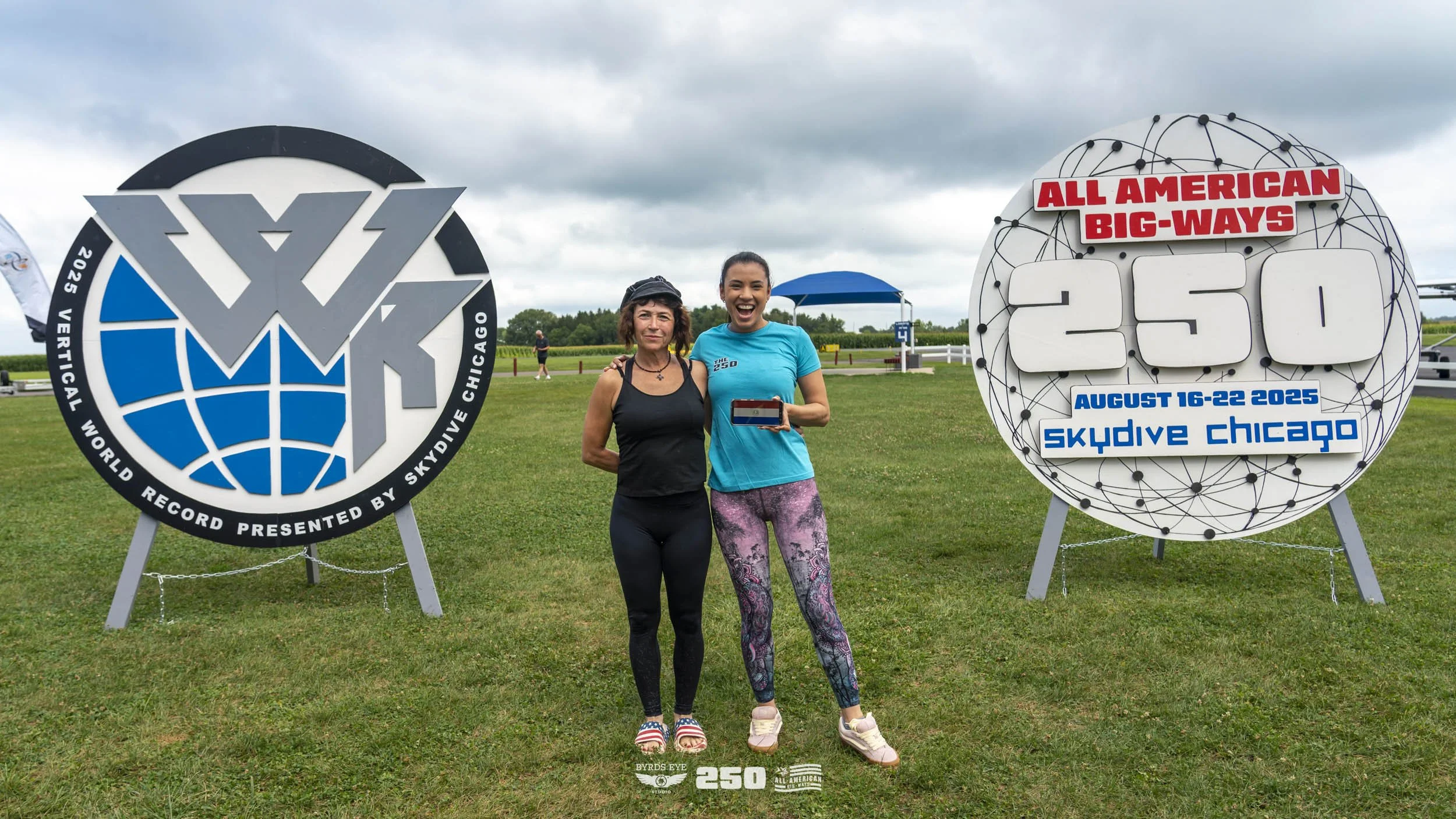 Two women standing on grass in front of large signs with event details. One woman is wearing a black tank top, leggings, and sandals, while the other woman is wearing a blue T-shirt and colorful leggings, holding a small flag. The signs indicate a re