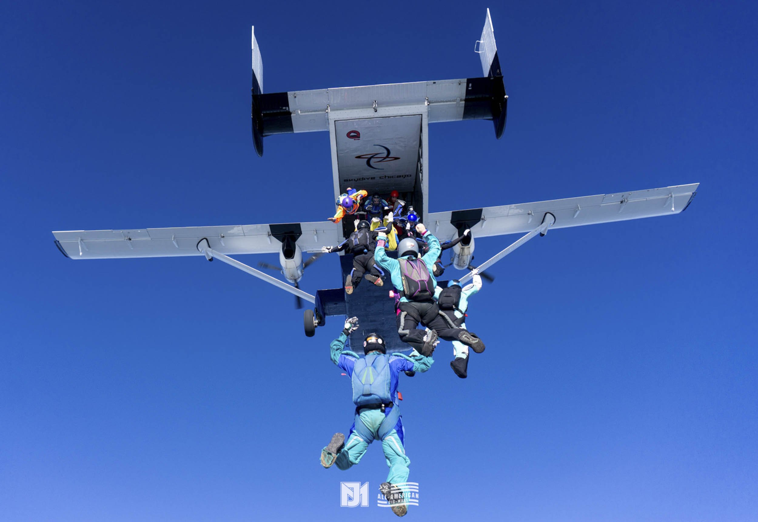 Skydivers free-falling from an airplane in clear blue sky, mid-descent, with some skydivers holding onto each other.