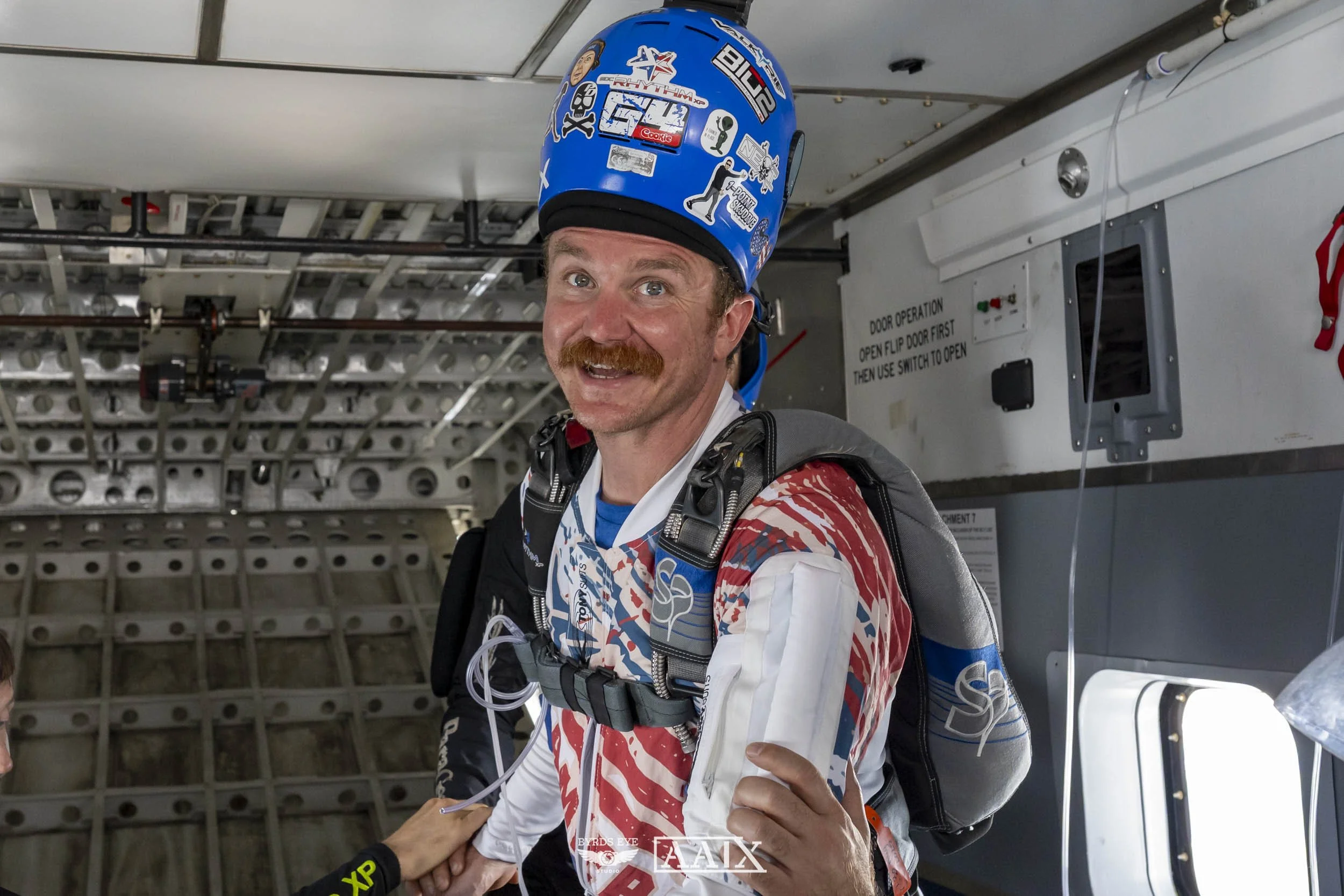 A man wearing a blue helmet with stickers, a red, white, and blue racing suit, and a parachute harness inside an aircraft, preparing for a skydive.