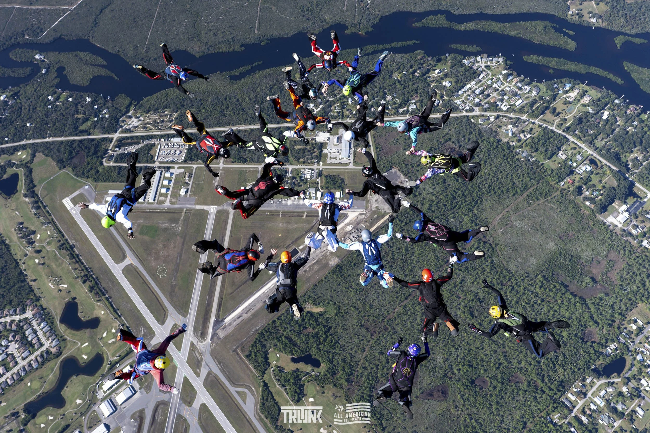 A group of skydivers in colorful jumpsuits and helmets, holding hands and forming a circle, free-falling over a green landscape with roads and bodies of water below.
