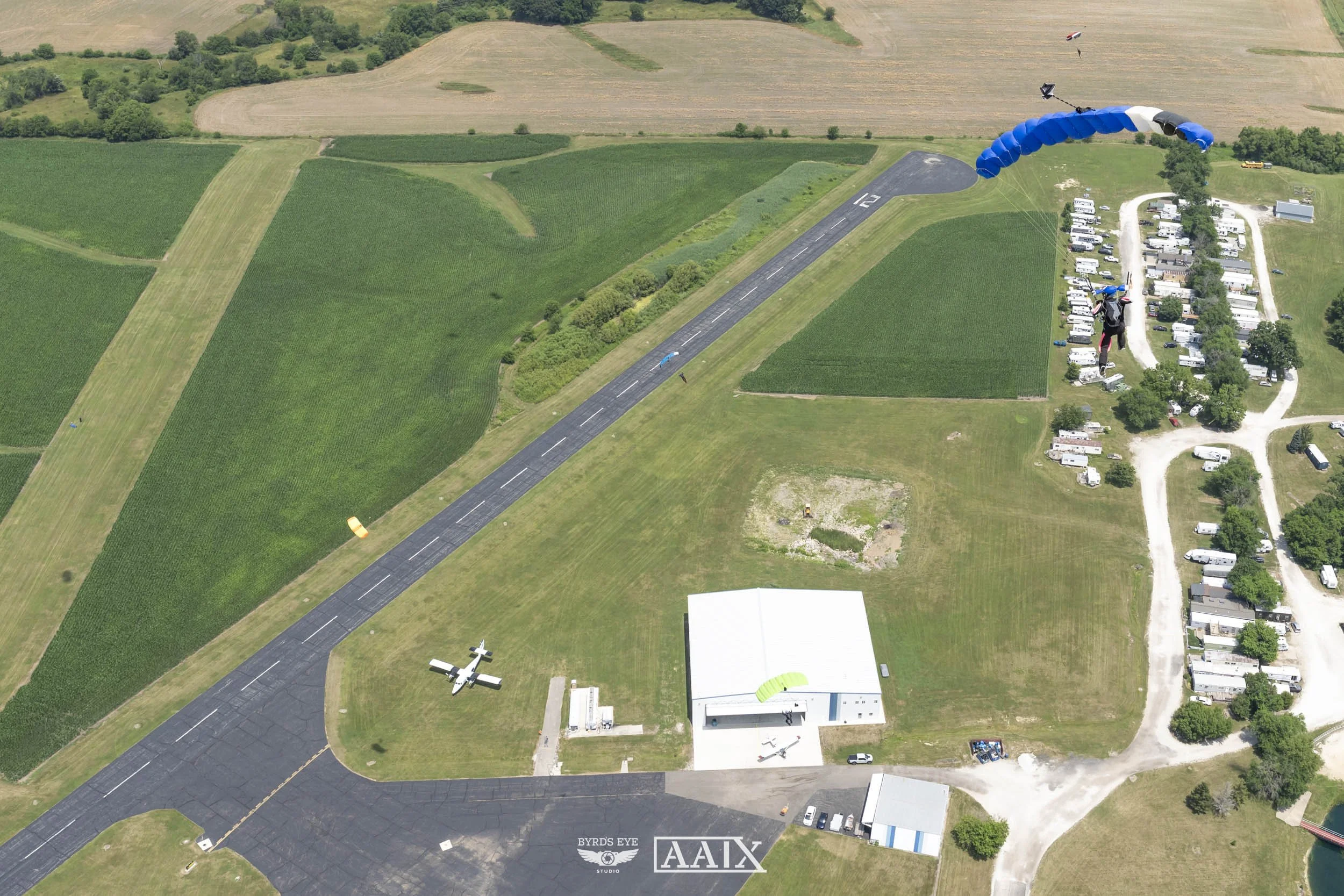 An aerial view of a small airport with a runway, a small airplane parked on the tarmac, and two skydivers with parachutes descending towards the ground.