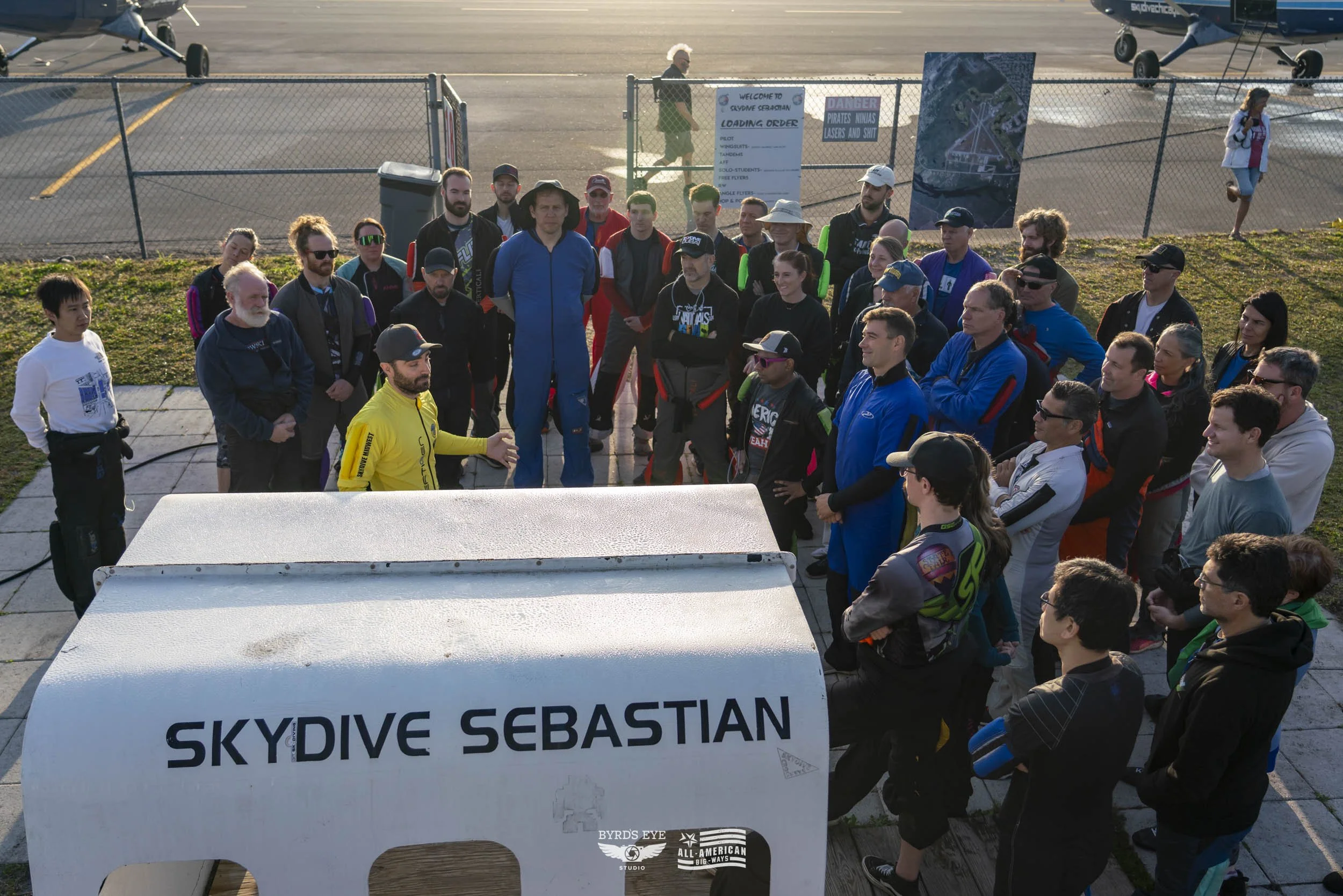 Group of people at Skydive Sebastian airport, listening to instructor before a jump, with small aircraft in background and informational signs, fencing, and parked planes visible.