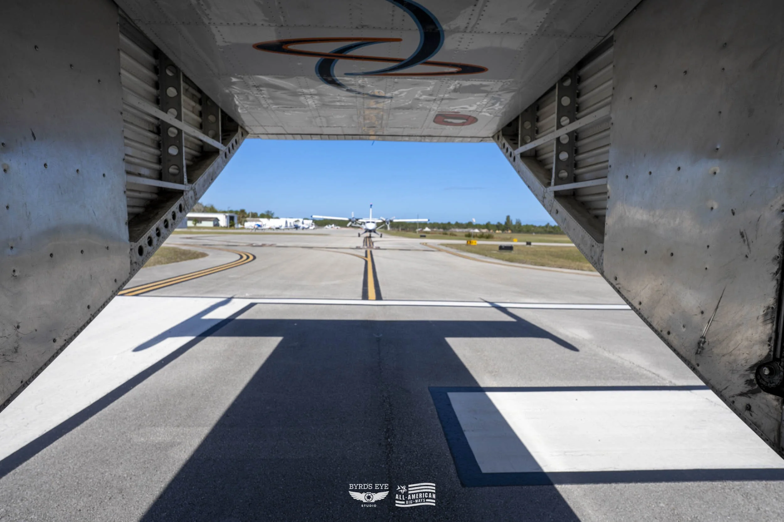 View through aircraft cargo door showing parked plane on tarmac with clear blue sky, several other planes, and airport buildings in the background.