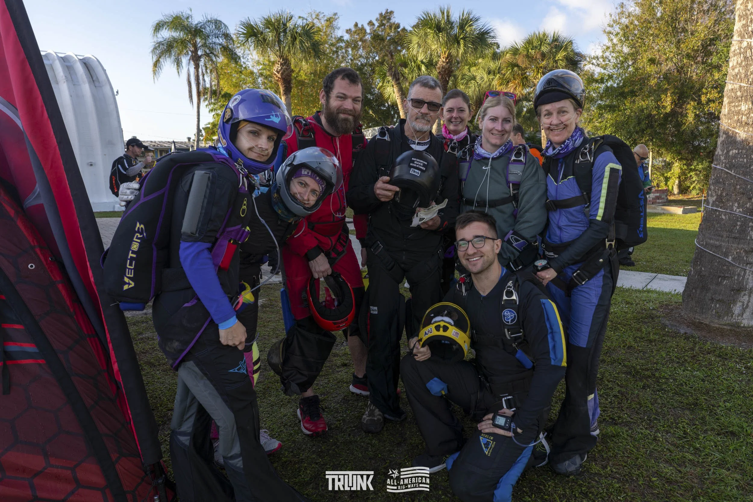 Group of skydivers wearing jumpsuits, helmets, and harnesses smiling together outdoors on a grassy area with palm trees in the background.