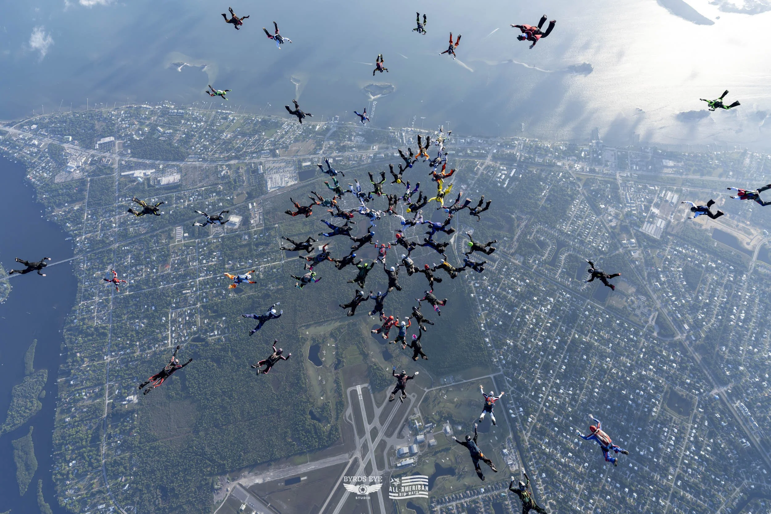 A large group of skydivers in formation above a city, with a parachute runway visible on the ground.
