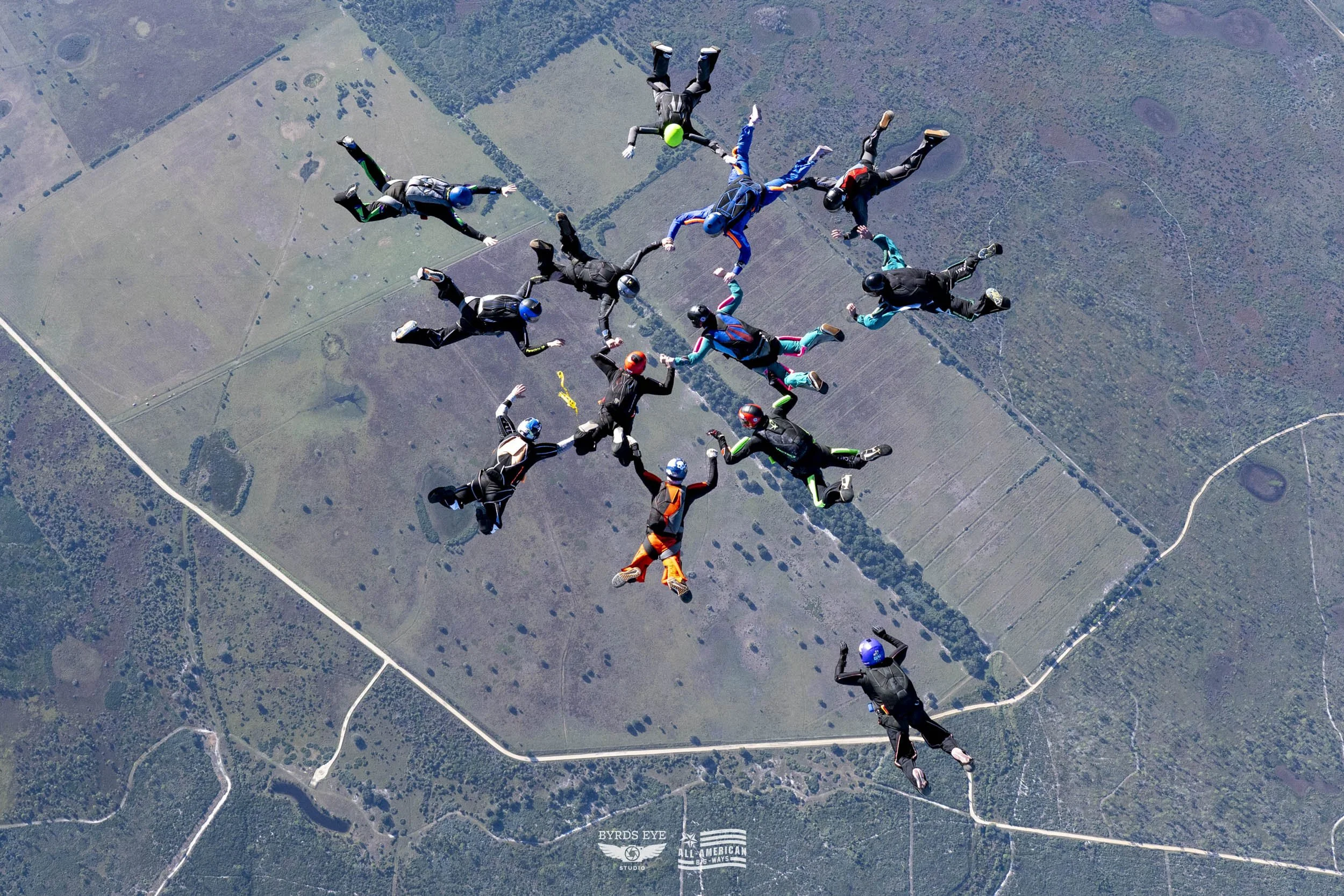 Group of skydivers in freefall formation holding hands forming a star shape over farmland and forested area.