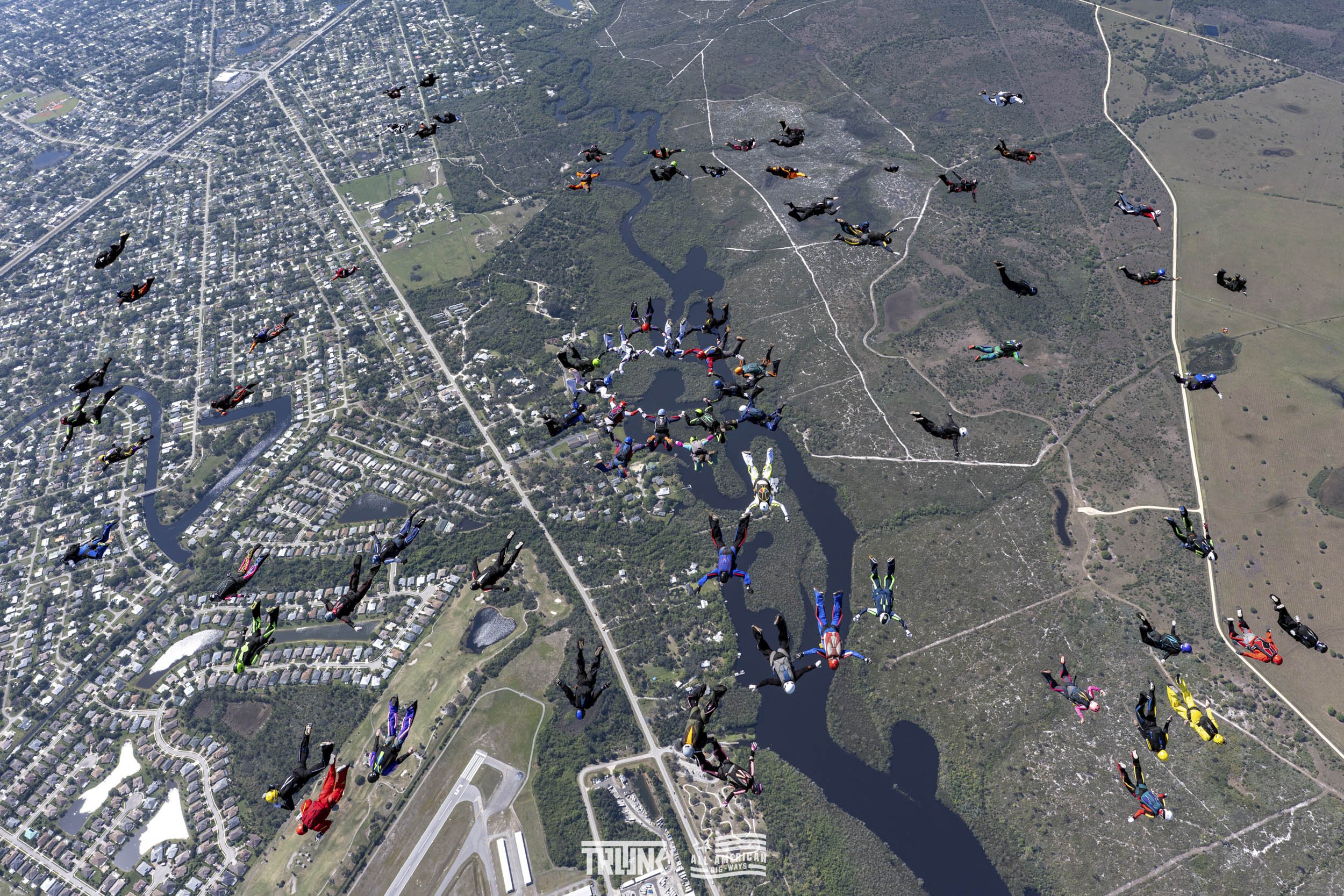 Multiple skydivers in free fall over a landscape that includes a river, roads, residential areas, and green fields.