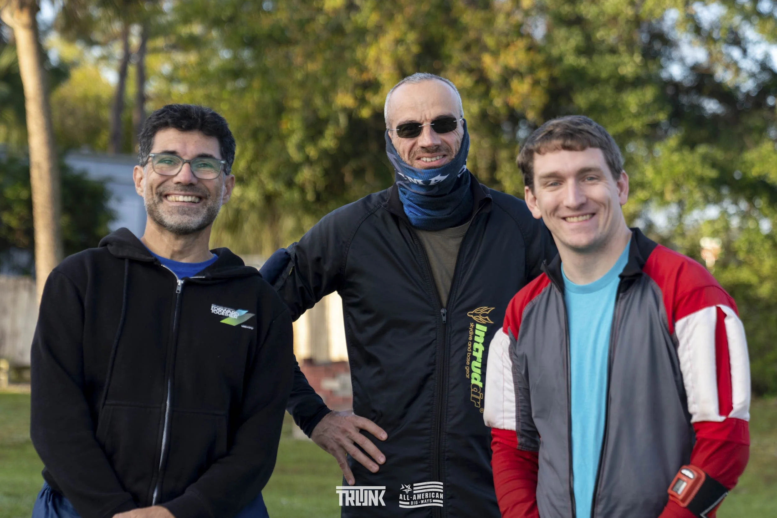 Three men standing outdoors in a park, smiling at the camera. The man on the left has dark hair, glasses, and a beard, wearing a black jacket. The man in the middle is bald, wearing sunglasses, a black jacket, and a bandana around his neck. The man o