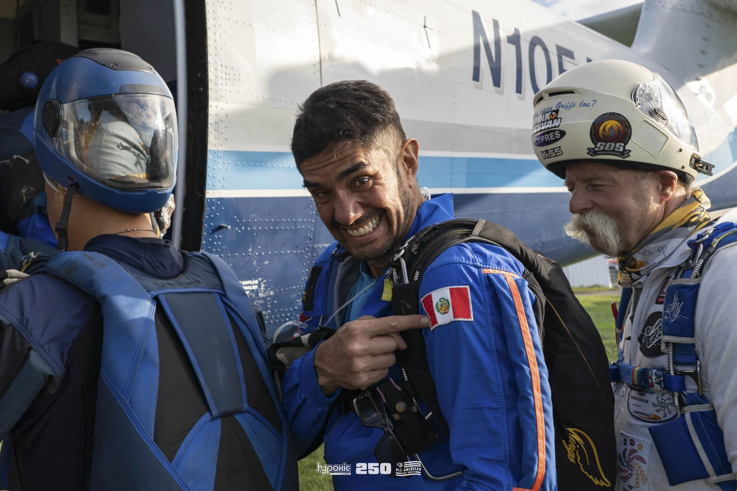 Three skydivers preparing for a jump, with one pointing to his Peruvian flag patch on his blue jumpsuit, standing next to a small airplane.