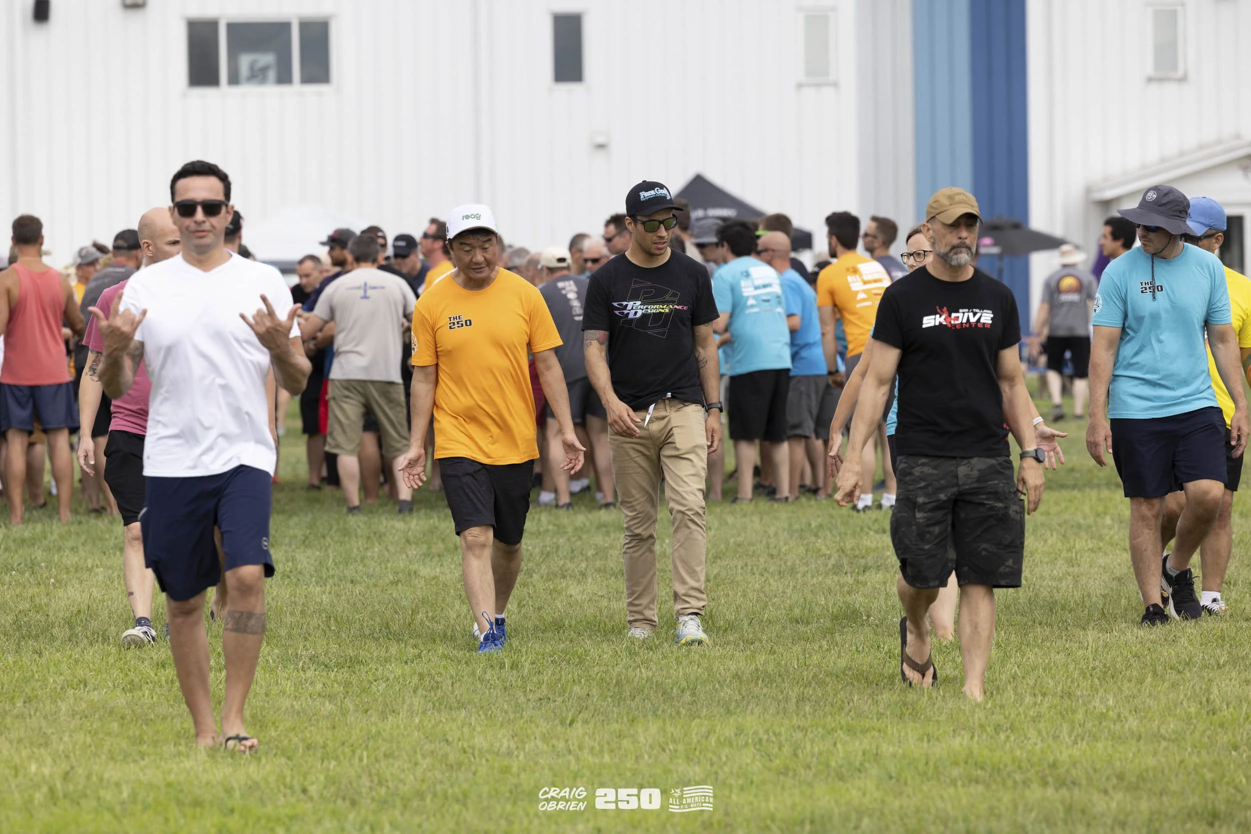 Group of diverse men walking on grass during outdoor event with a crowd in the background.