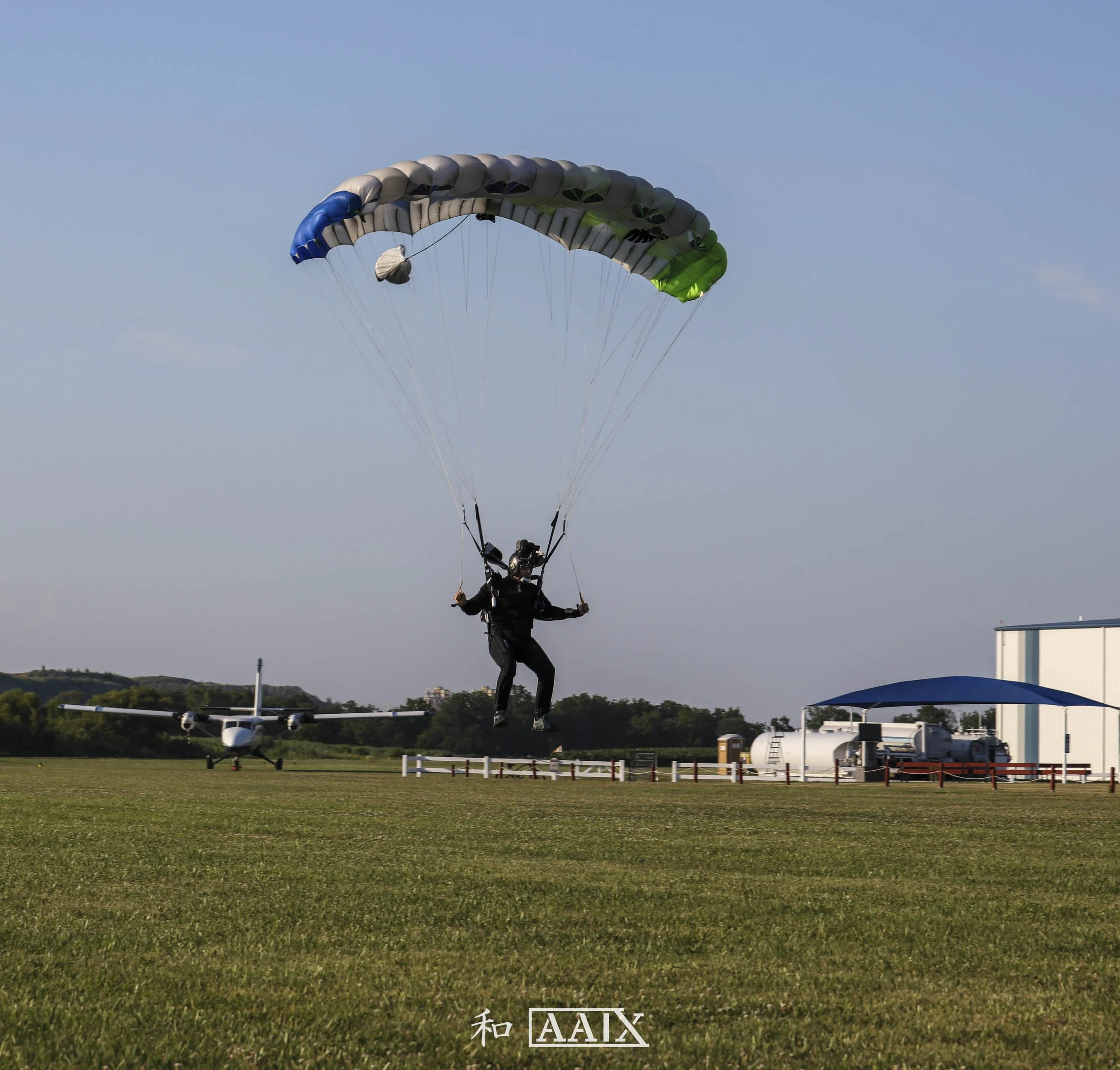 Person skydiving with a parachute on a grassy field with small airport buildings and airplane in the background.