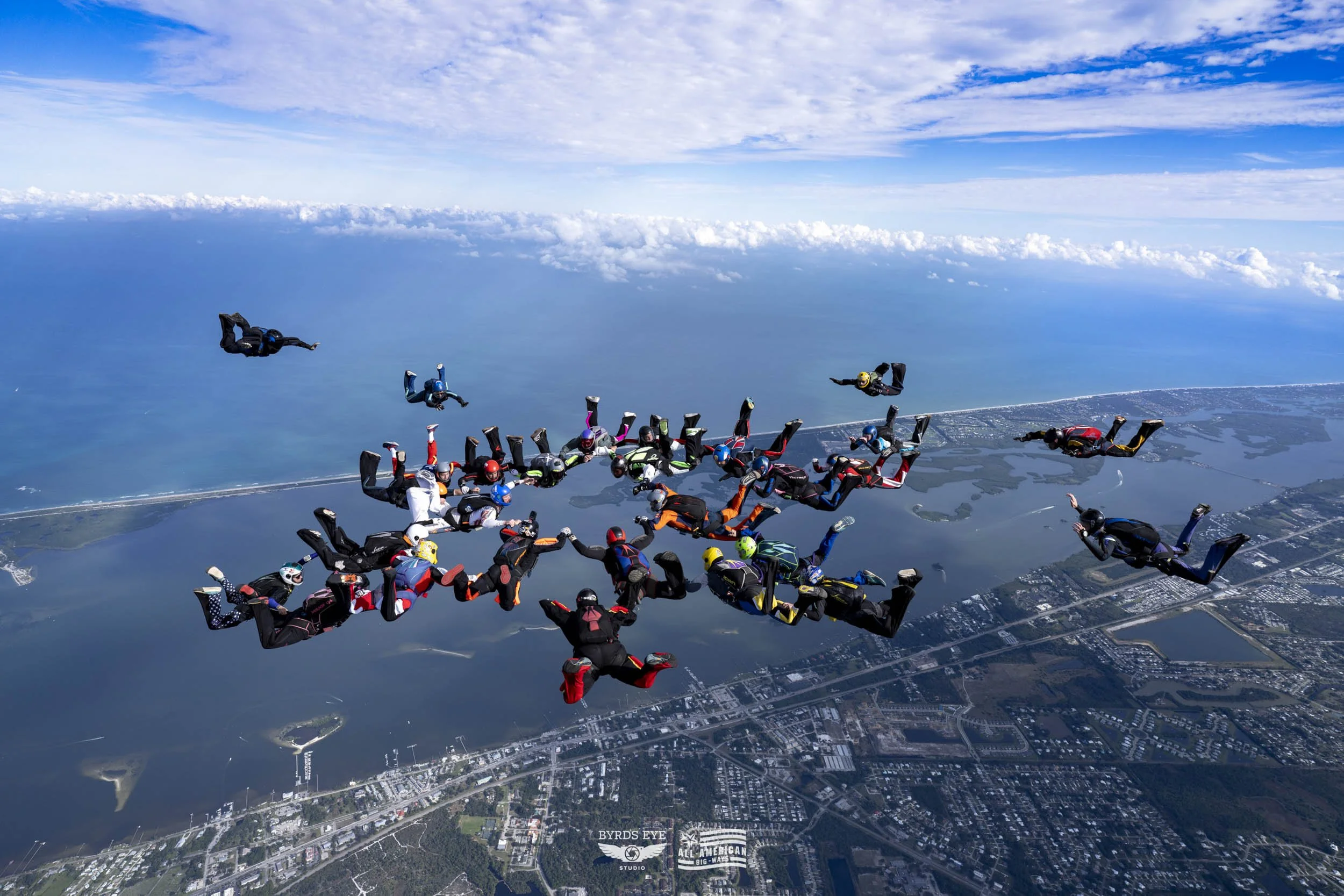 Group of skydivers in freefall formation over a landscape with water and land, under a partly cloudy sky.