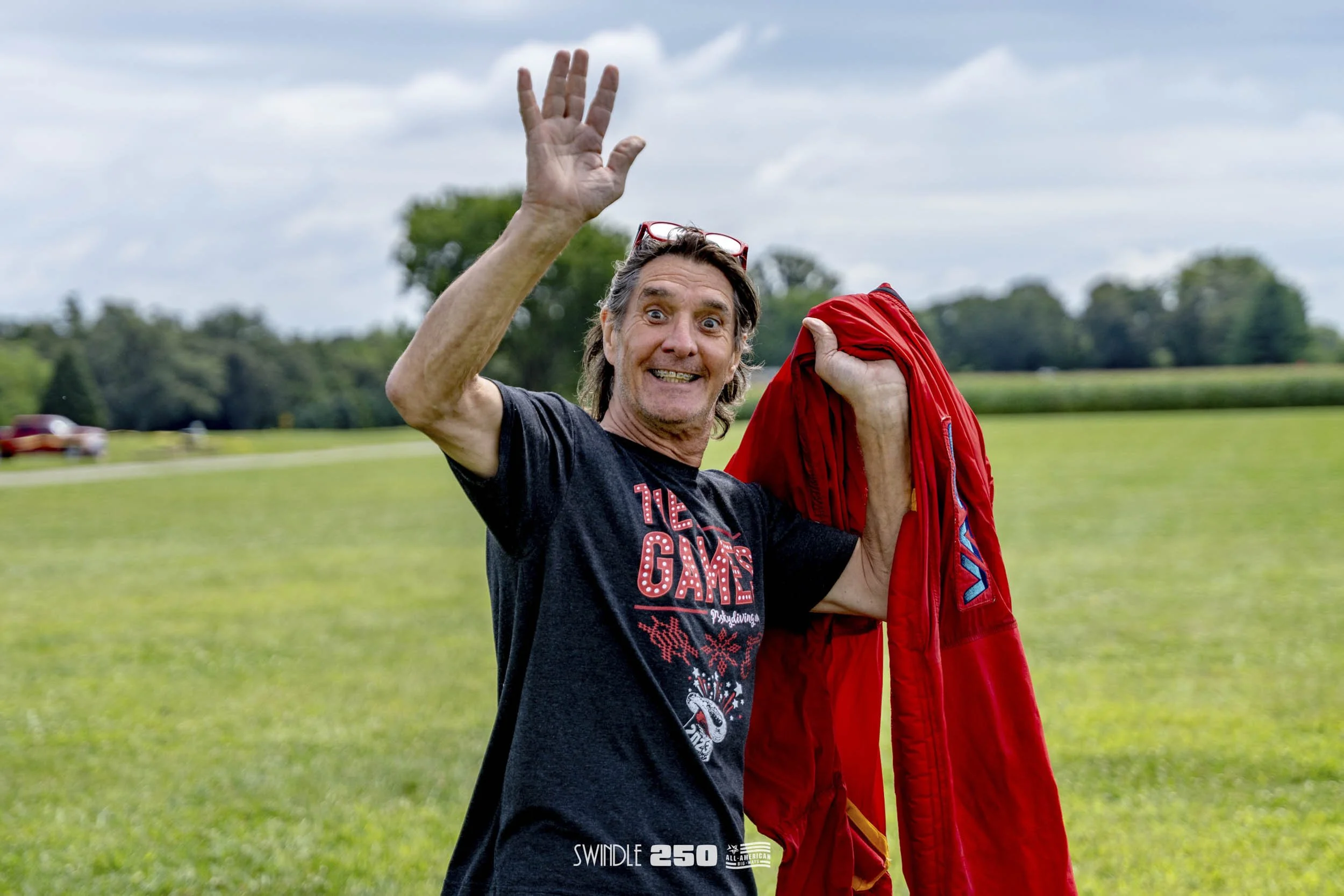 A man outdoors in a grassy field, waving and smiling, holding a red jacket over his shoulder, wearing glasses on his head, and a black T-shirt with red and white graphics, with trees and cars in the background.