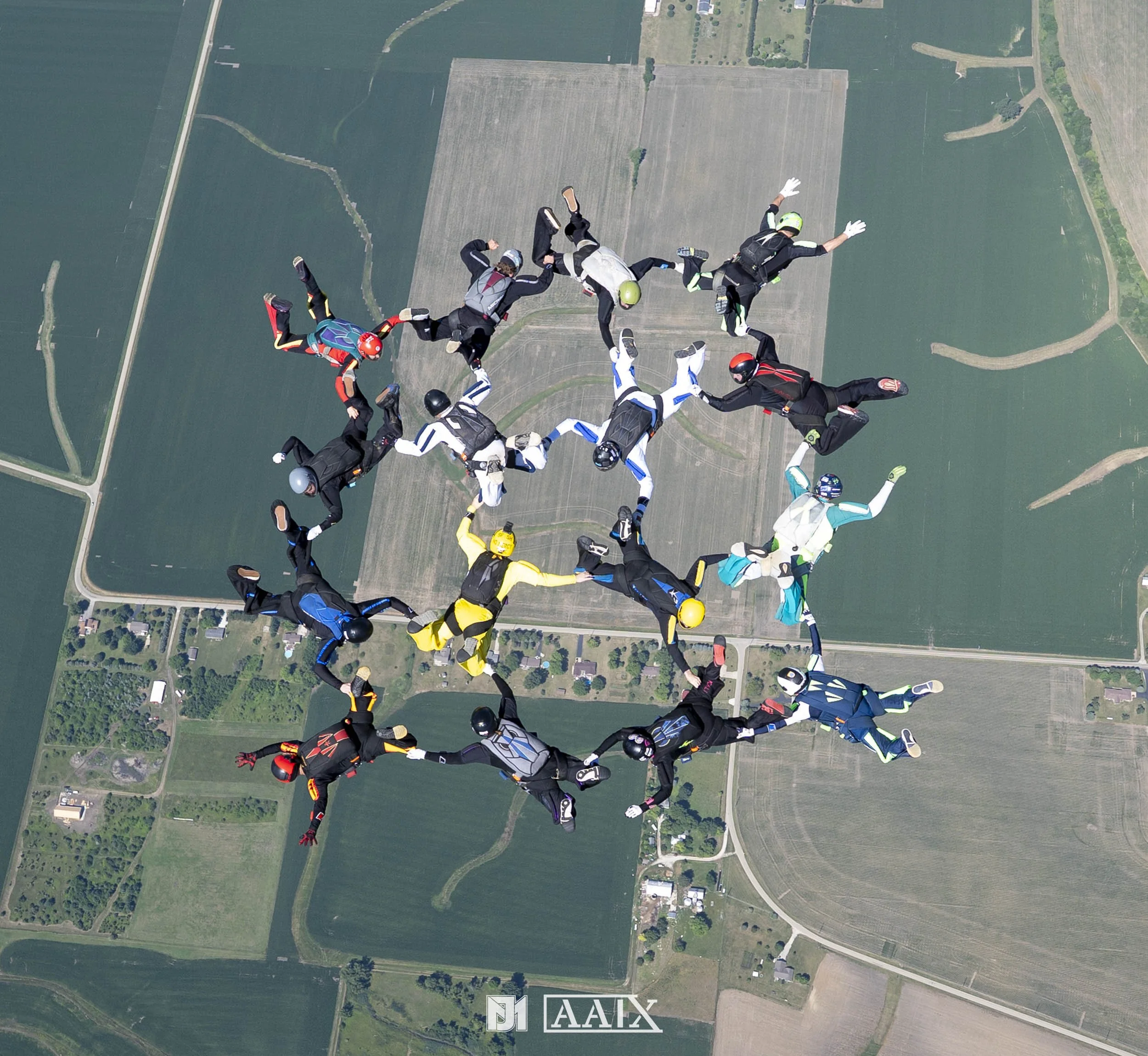 Skydivers forming a star shape while free-falling over farmland, wearing colorful jumpsuits and helmets.