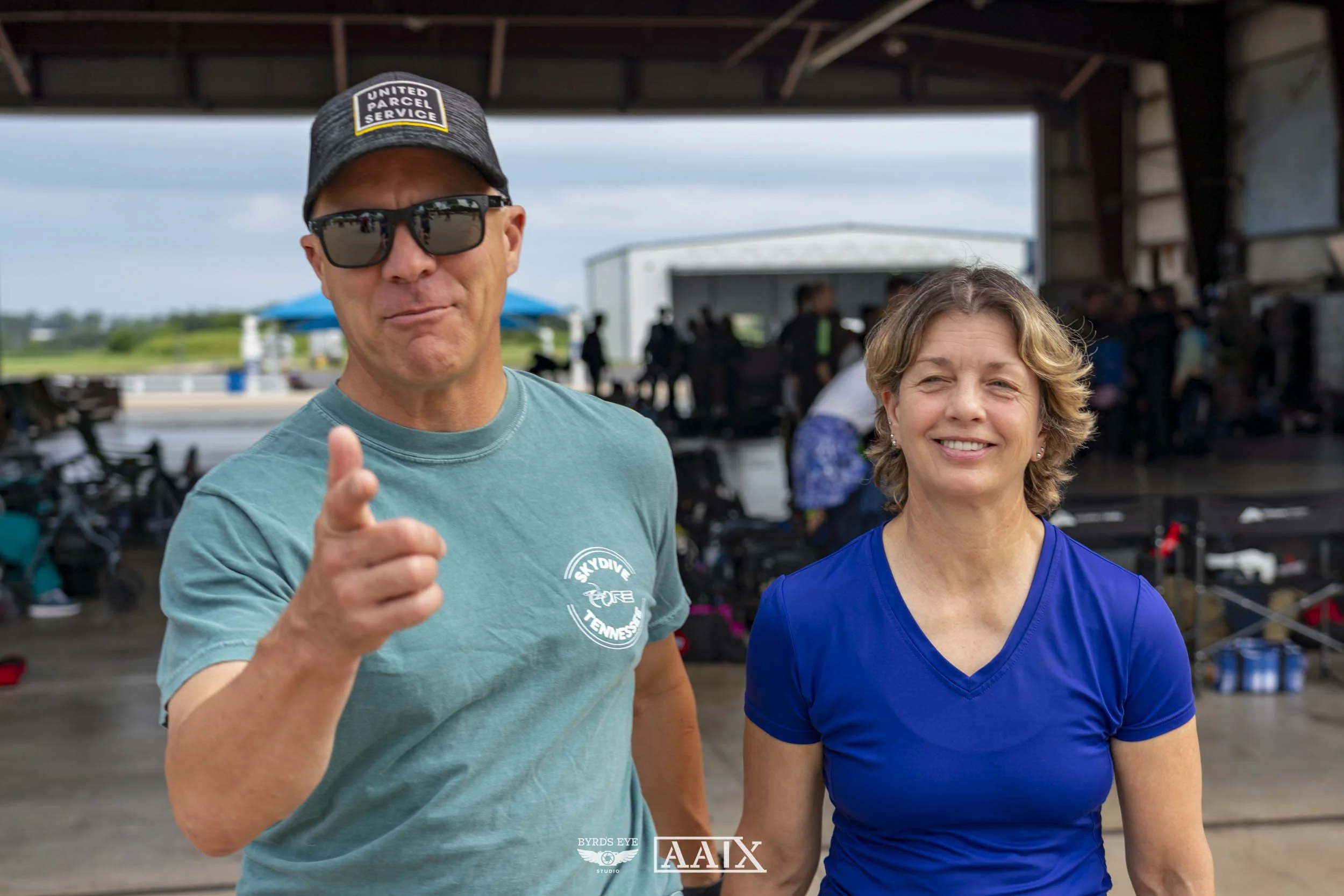 A man wearing sunglasses, a gray baseball cap with 'United Parcel Service' written on it, and a teal t-shirt pointing toward the camera, standing next to a woman with short blond hair wearing a blue shirt, at an outdoor event under a pavilion with pe