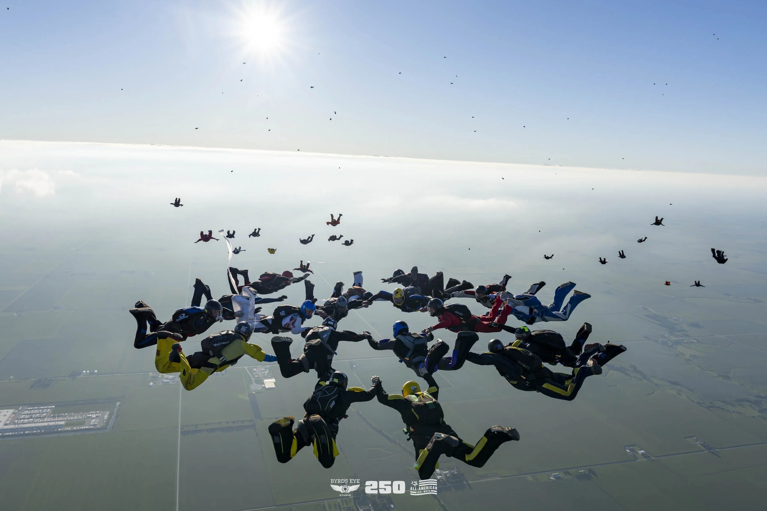 A group of skydivers holding hands forming a circle in free fall above the countryside, with others falling in the background and the sun shining in the sky.