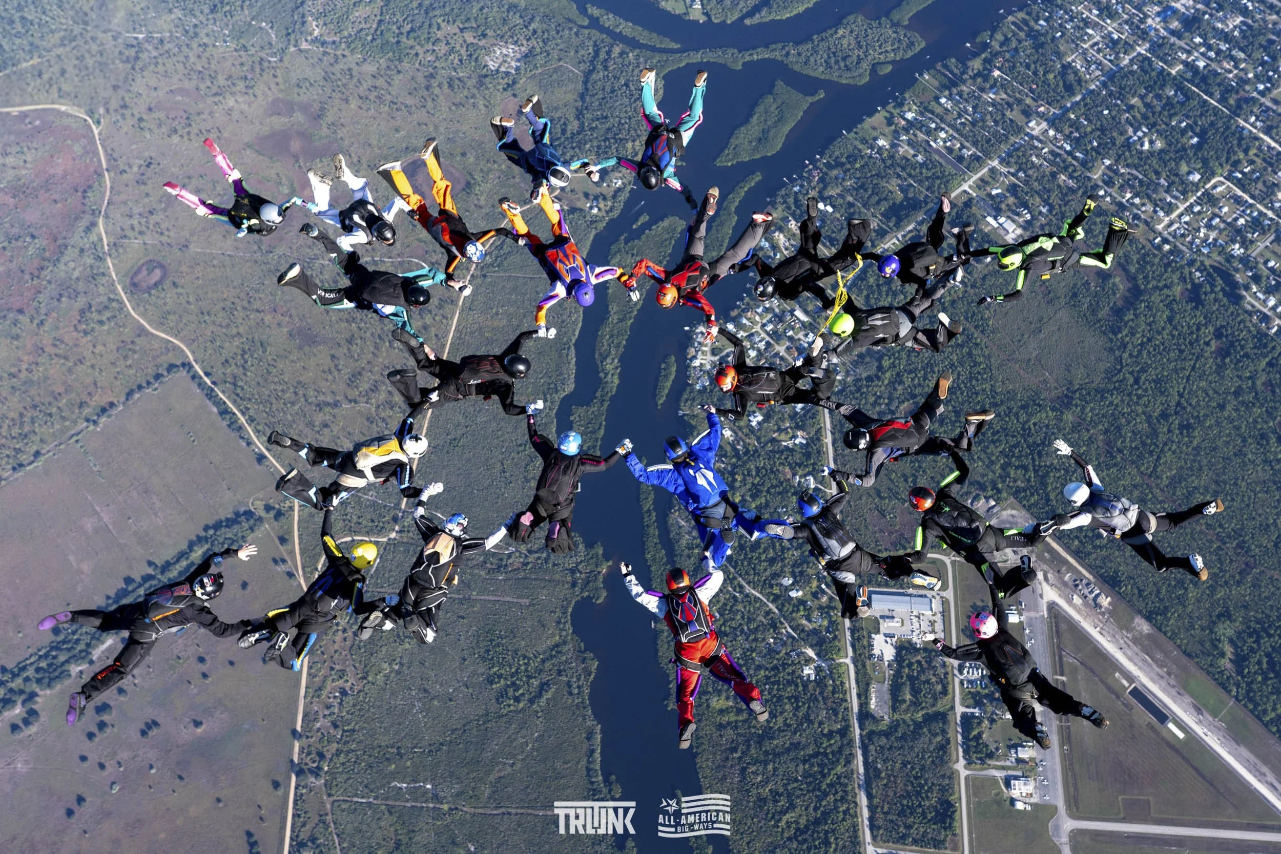 Group of skydivers forming a star pattern while free-falling over a landscape with water, trees, and fields.